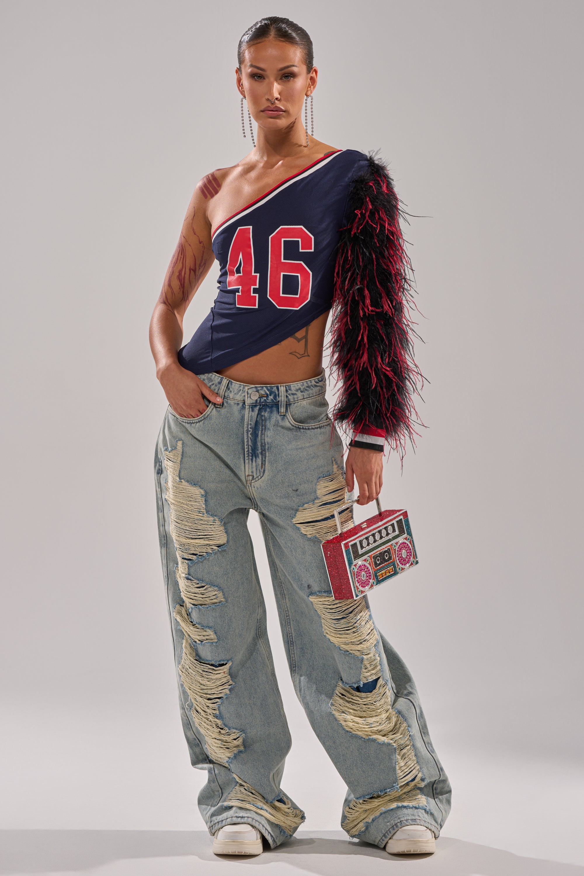 Wearing the SUPERBOWL PARTY ONE SLEEVE TEE with feather accents and "46," she pairs ripped baggy jeans and white sneakers, confidently posing with a boombox-shaped purse against a plain background. 5