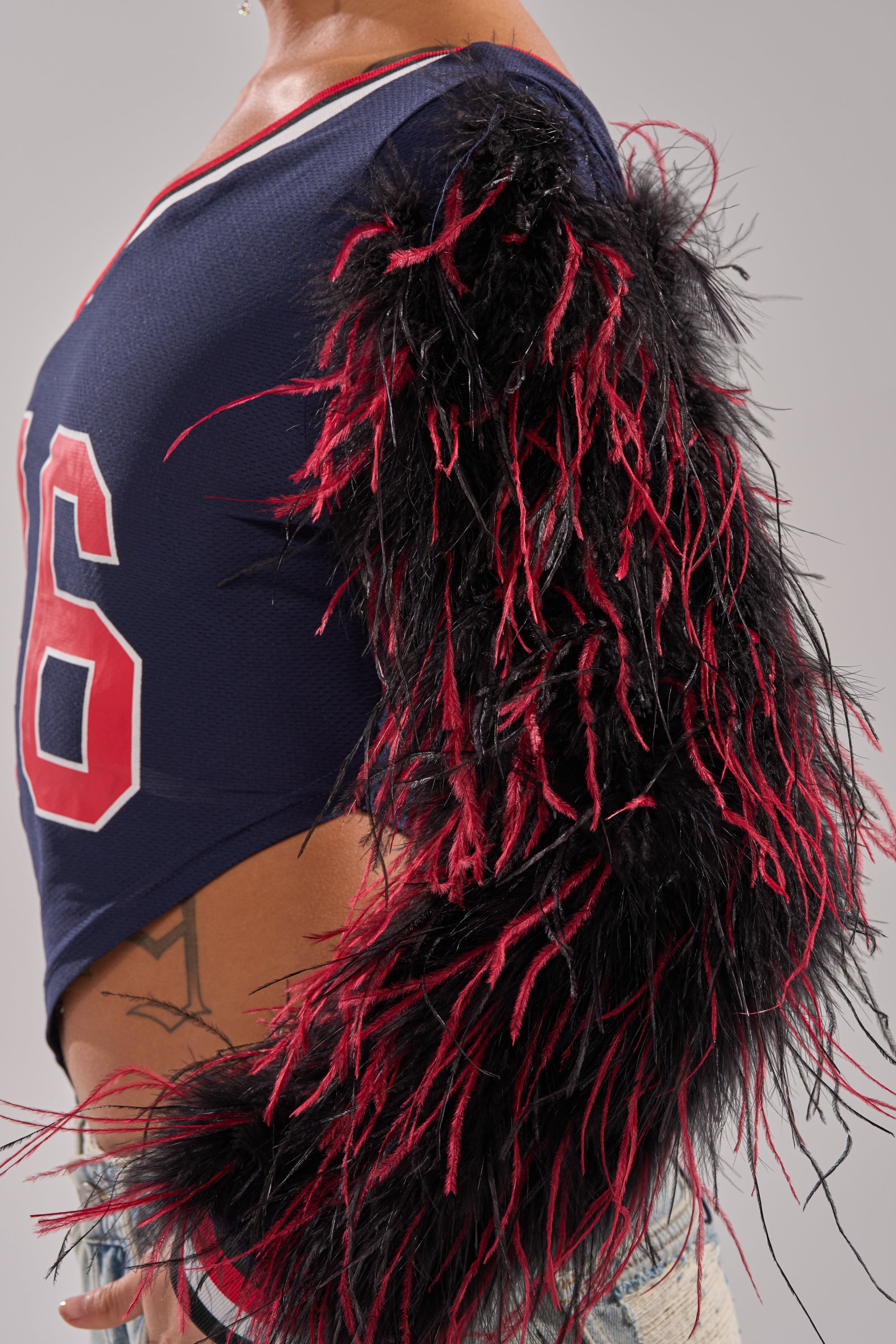 A person wears the blue SUPERBOWL PARTY ONE SLEEVE TEE featuring the number 16, an oversized sleeve with black and red feathers, and displays a tattoo near their waist above ripped denim shorts. 7