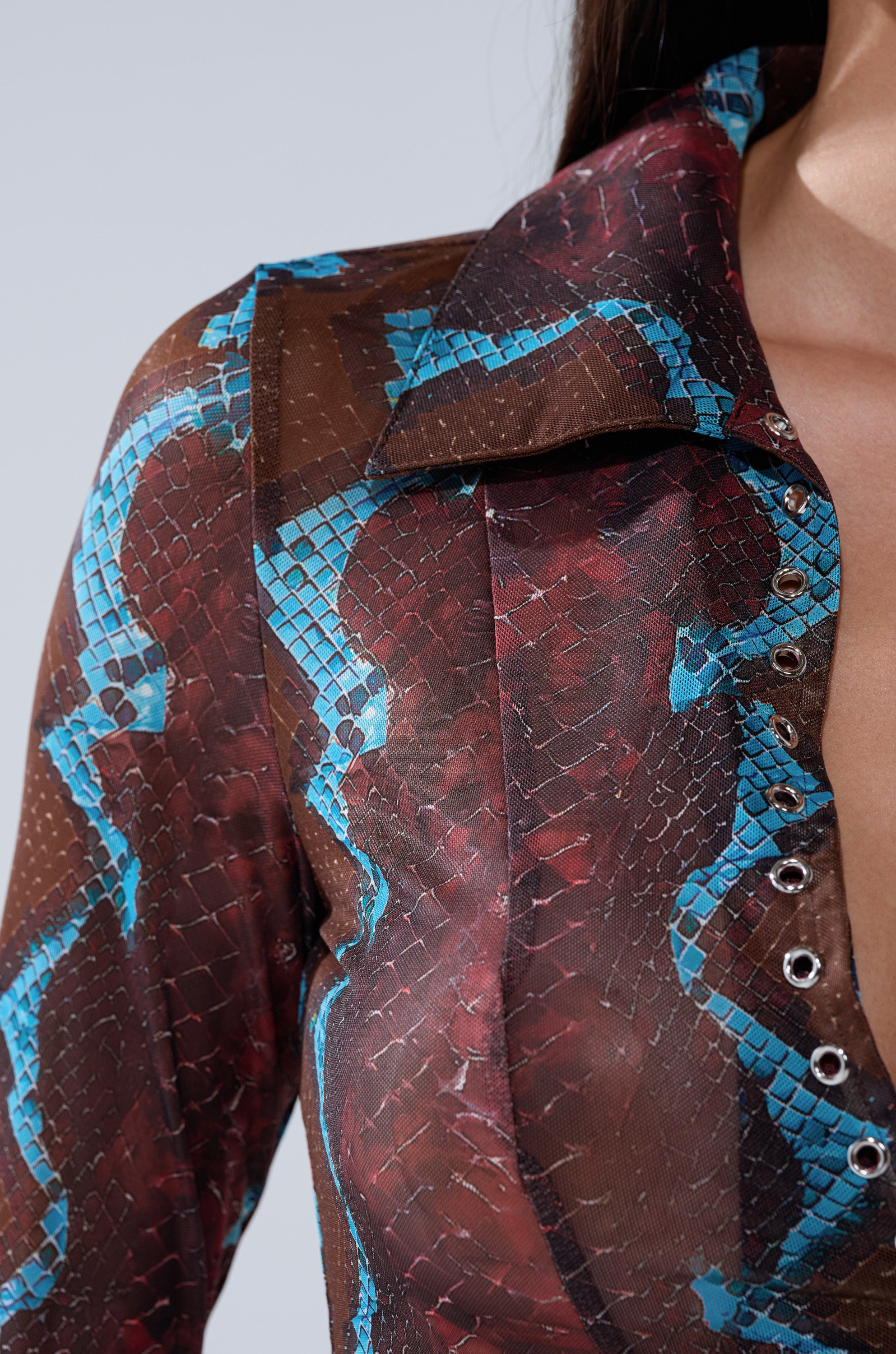 A close-up of the DONT BE A SNAKE BUTTON DOWN, featuring a shiny brown and blue snakeskin pattern and large collar, worn by someone with long dark hair. Only the mesh shirt and part of their neck are visible.