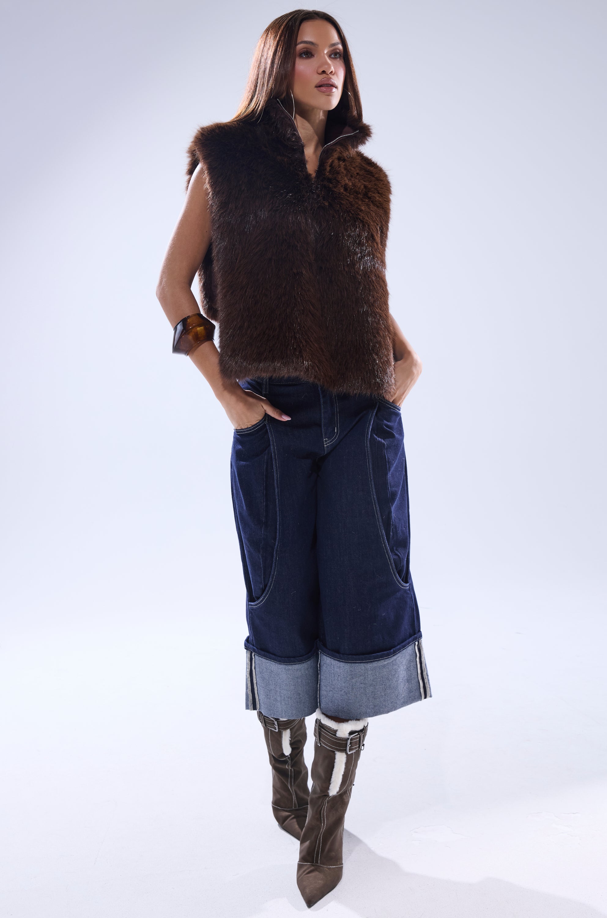 A woman stands confidently against a plain background in the SURE THANG FUR TURTLENECK PULLOVER, paired with dark wide-leg cuffed jeans, brown heeled boots, and a chunky bracelet, her hands tucked stylishly in her pockets. 6