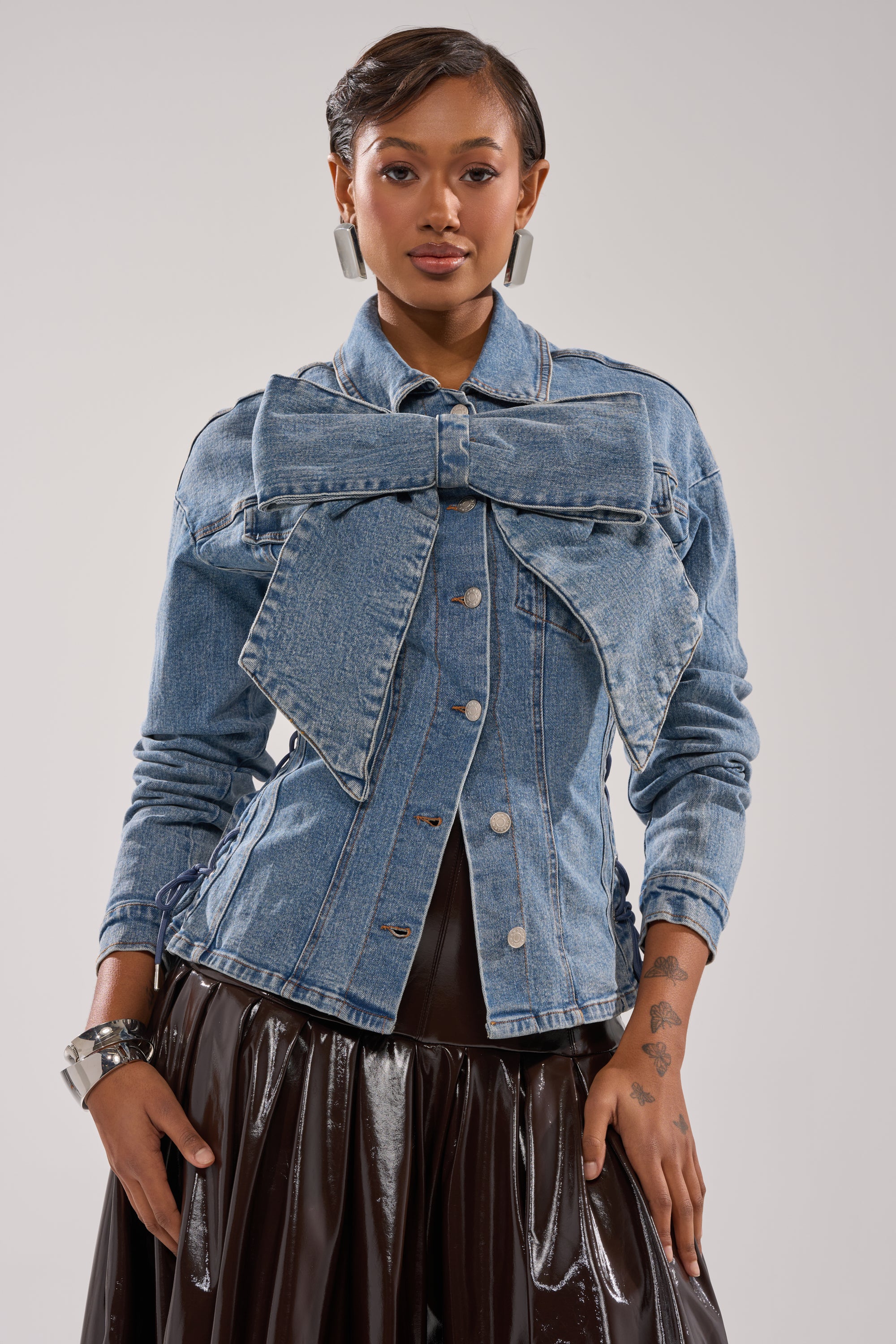 A woman wearing the DAY BREAK BUTTON DOWN BLOUSE, paired with large silver earrings and a black shiny skirt, stands facing forward against a plain light background.
