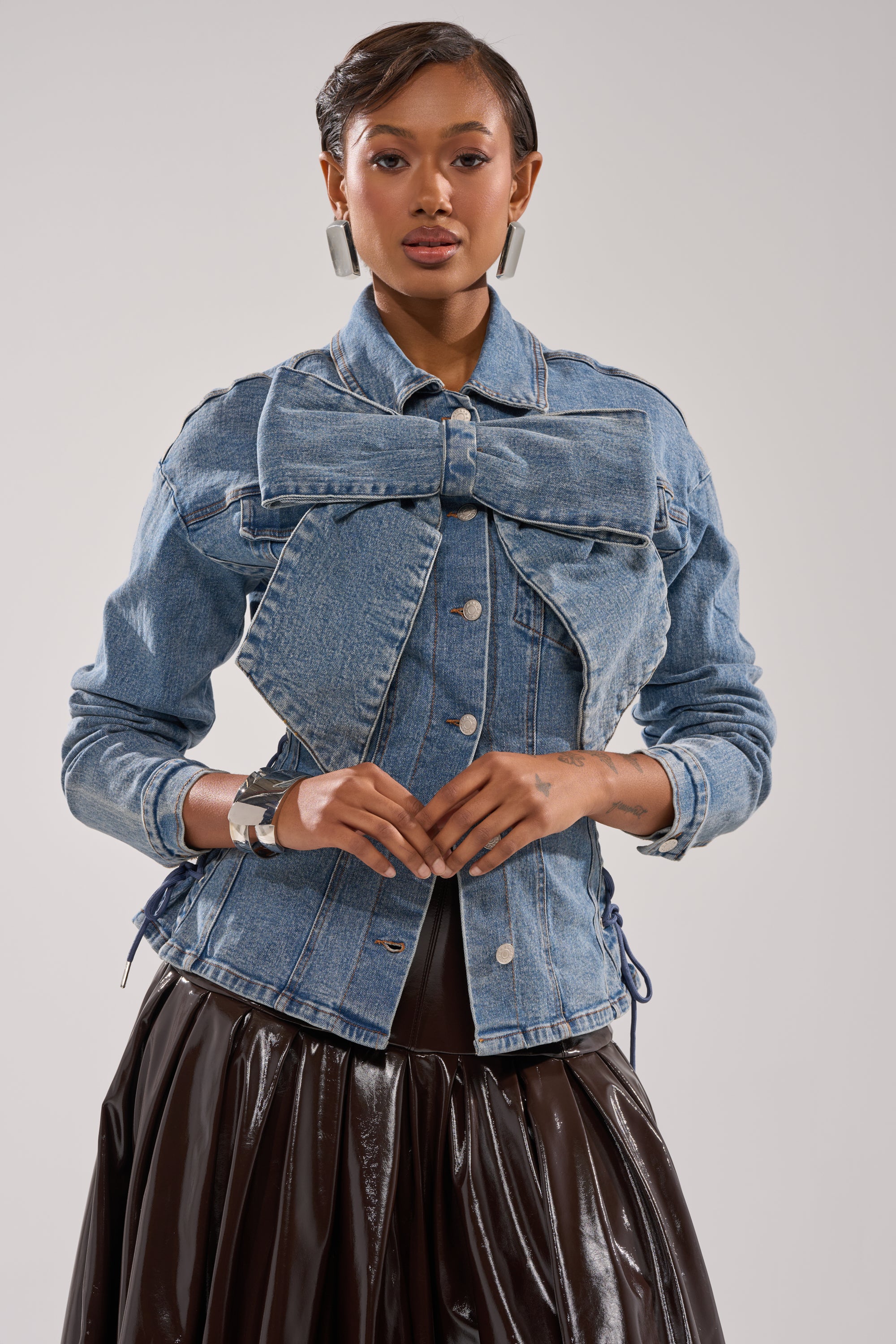 A woman poses in the DAY BREAK BUTTON DOWN BLOUSE with an oversized bow detail, paired with silver earrings, a chunky bracelet, and a dark pleated skirt. She stands against a plain, light-colored background.