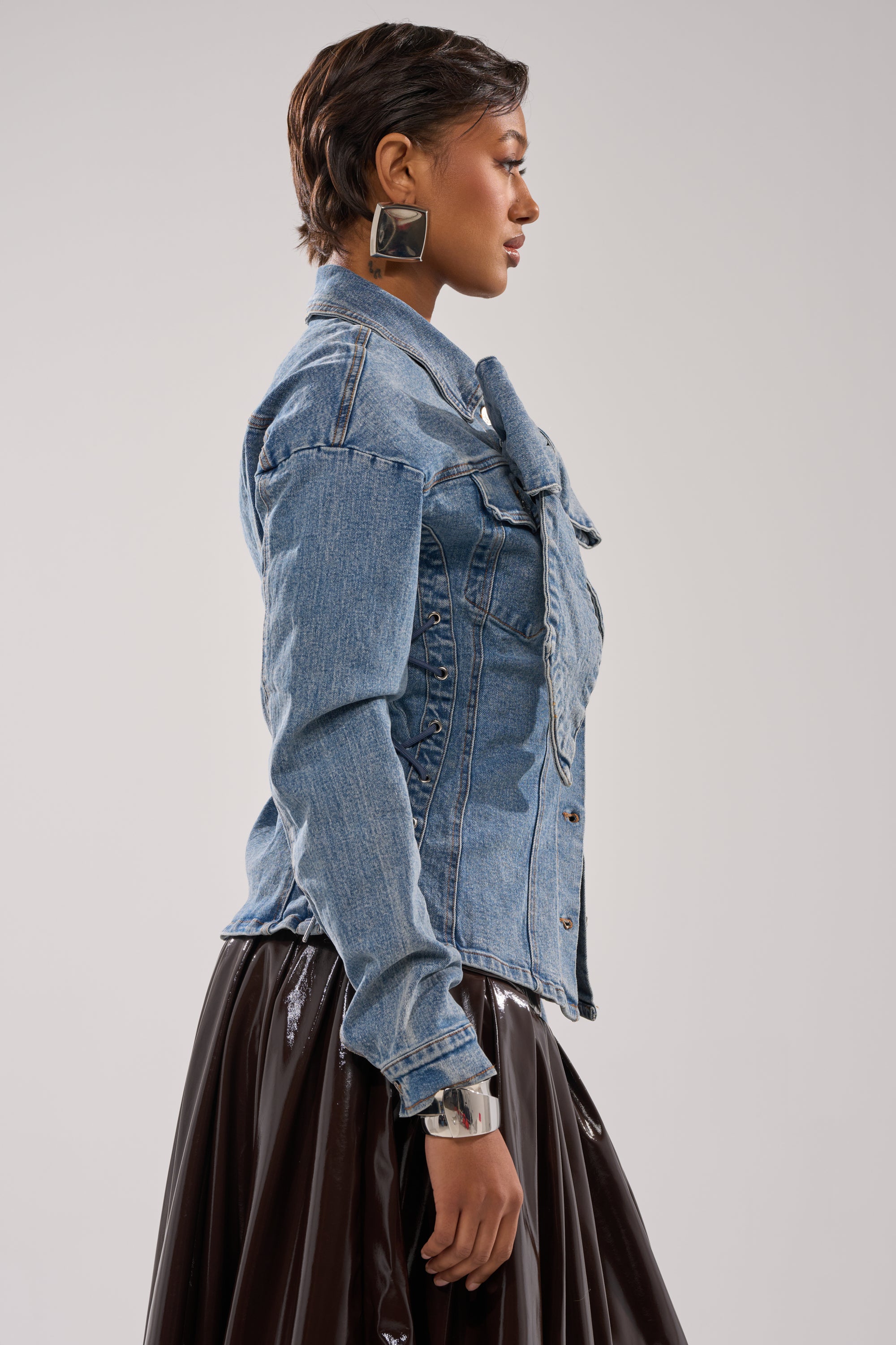 A woman stands in profile against a plain background, wearing the DAY BREAK BUTTON DOWN BLOUSE, paired with a black shiny skirt, large square earrings, and a chunky silver bracelet.
