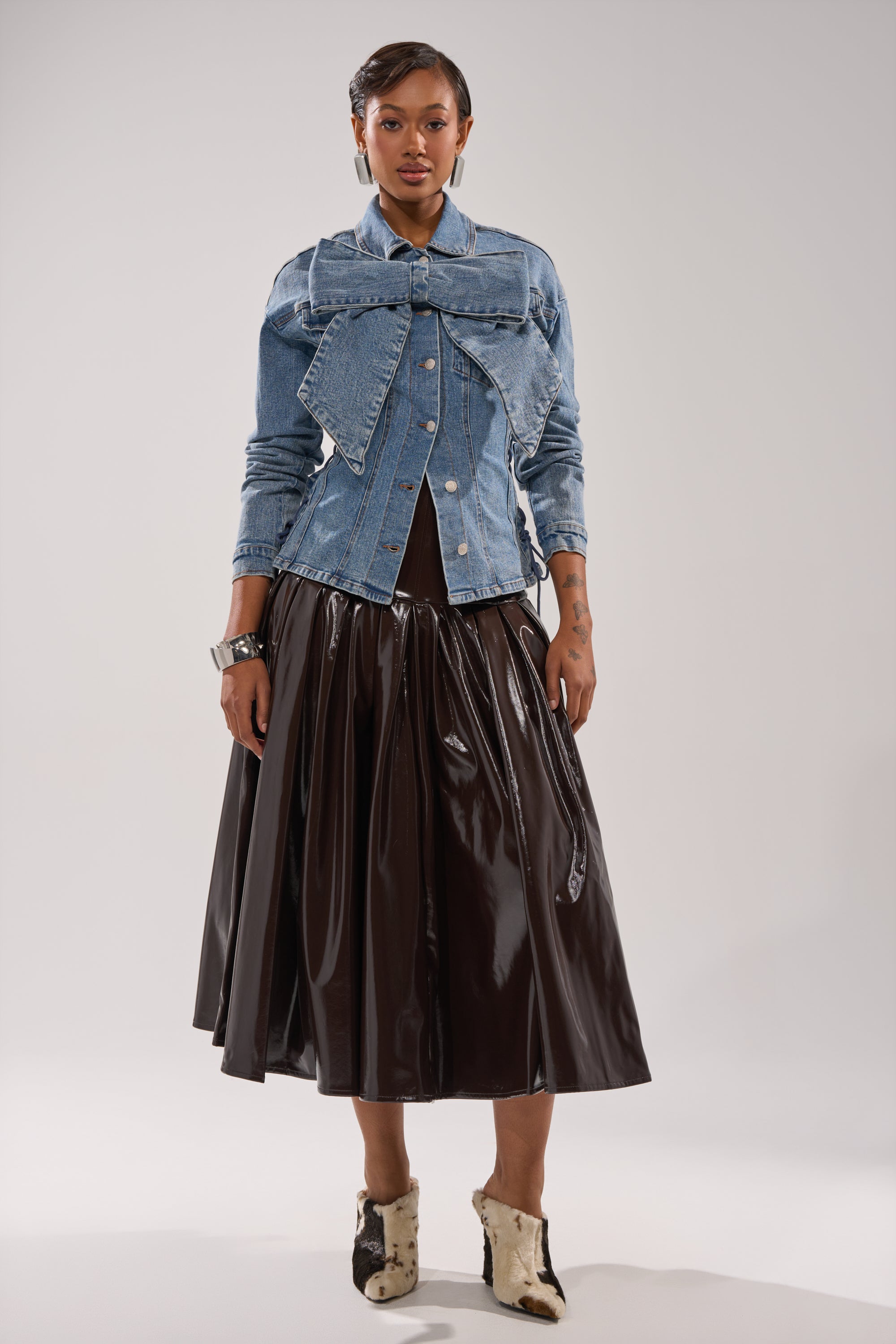A person stands confidently facing forward in the DAY BREAK BUTTON DOWN BLOUSE, paired with a shiny black midi skirt and ankle-high two-tone boots. The plain background highlights this stylish look.