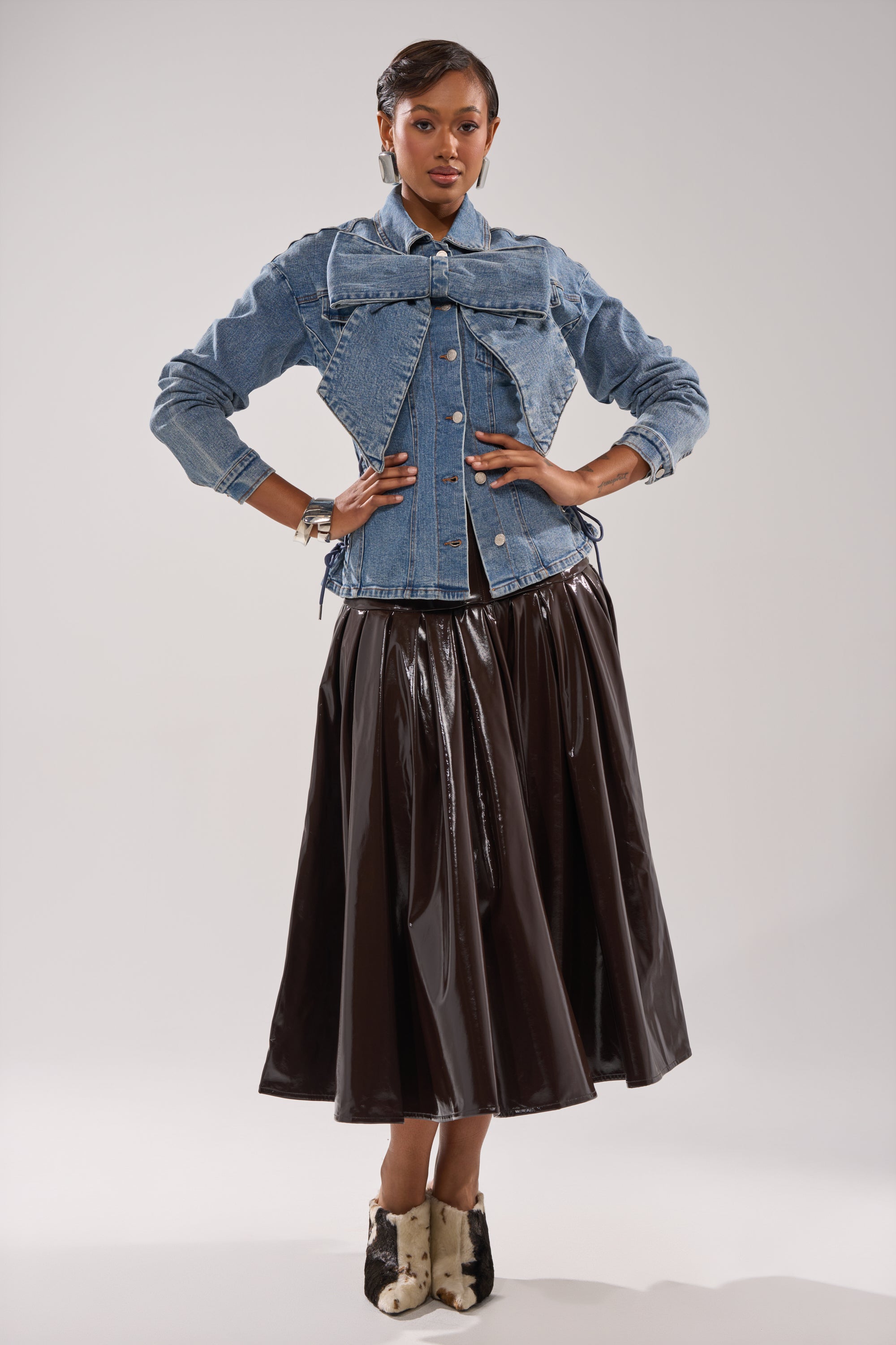 A person stands confidently in the DAY BREAK BUTTON DOWN BLOUSE, featuring a large collar, paired with a shiny black midi skirt and spotted ankle boots against a plain light background.
