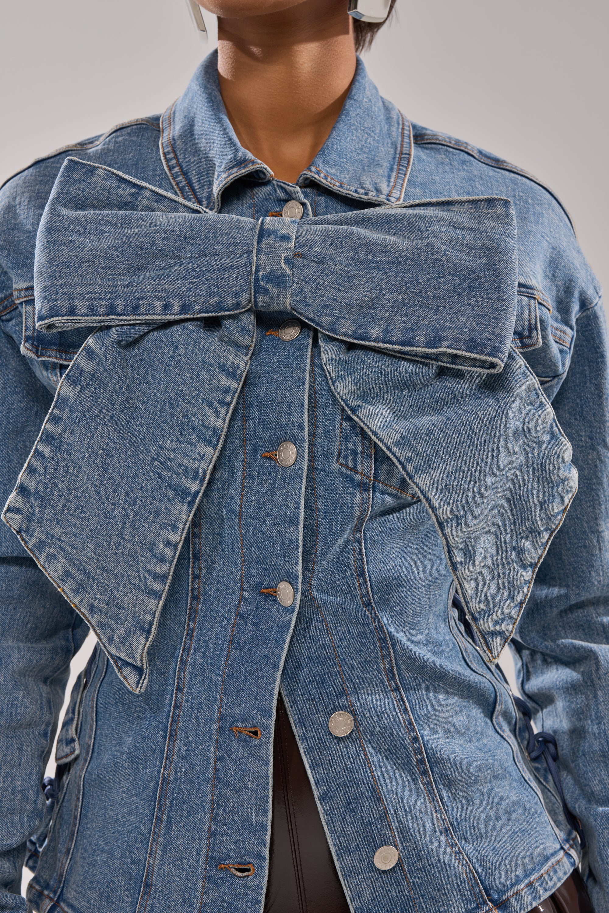 A person wearing the DAY BREAK BUTTON DOWN BLOUSE, a blue denim top with an oversized bow across the chest and silver buttons down the front.