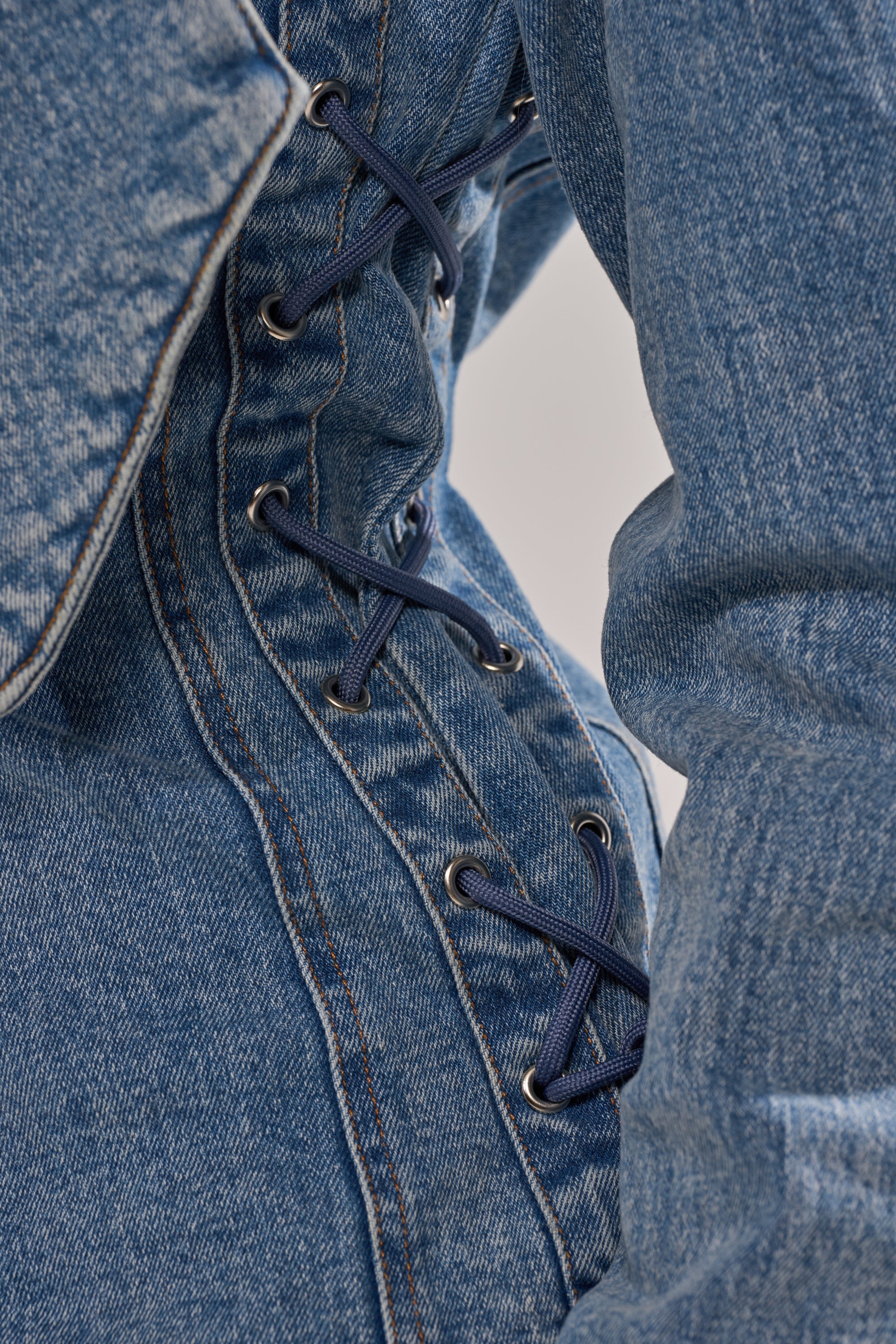 Close-up of the DAY BREAK BUTTON DOWN BLOUSE featuring blue denim fabric with dark blue laces threaded through metal eyelets in a crisscross, corset-style detail.