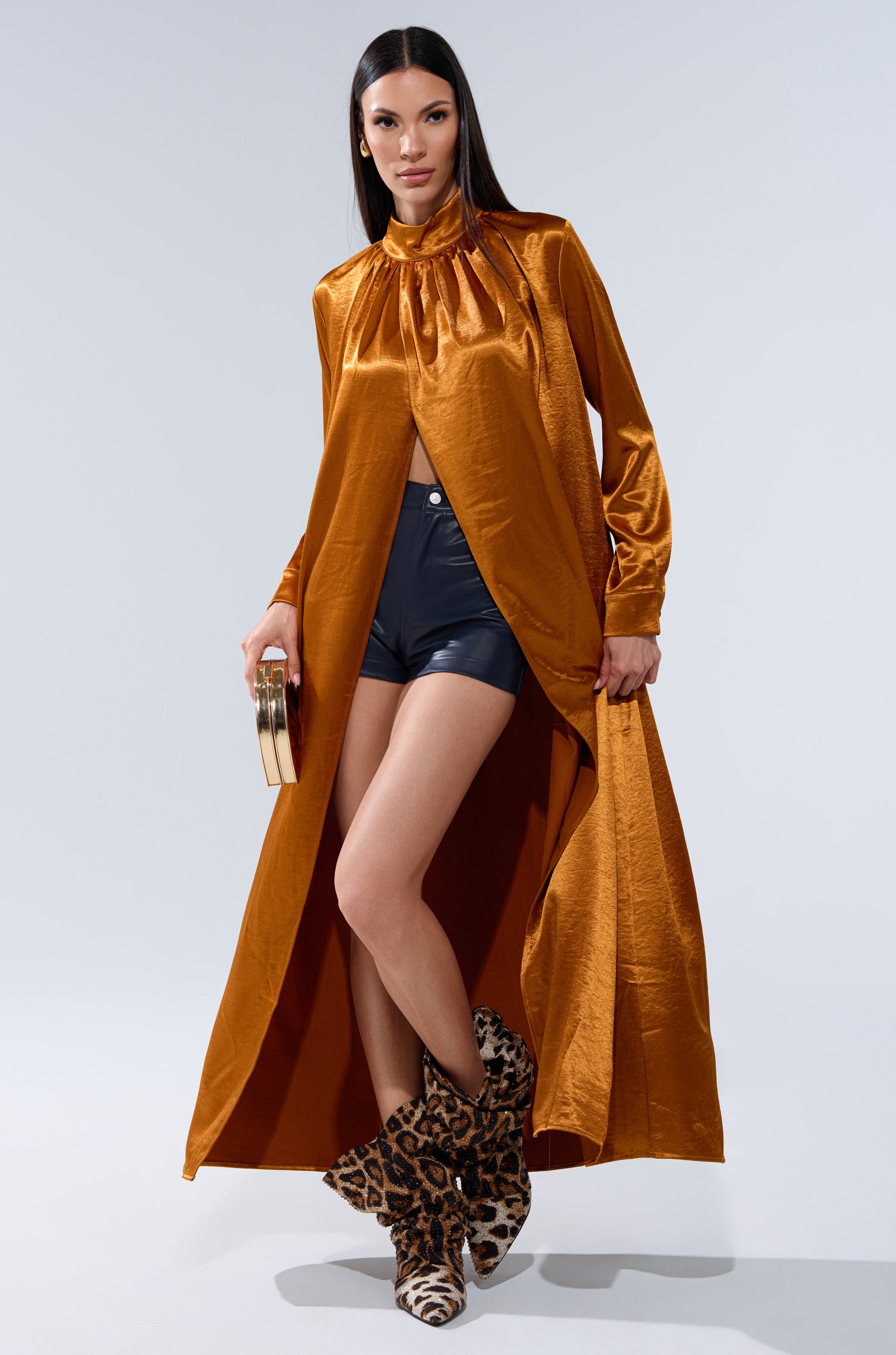 A woman models the QUEEN TYPE BLOUSE in shiny copper with a mock neckline and high slit, paired with black shorts, leopard print boots, and a round bag. She has long straight hair and stands against a plain light background.