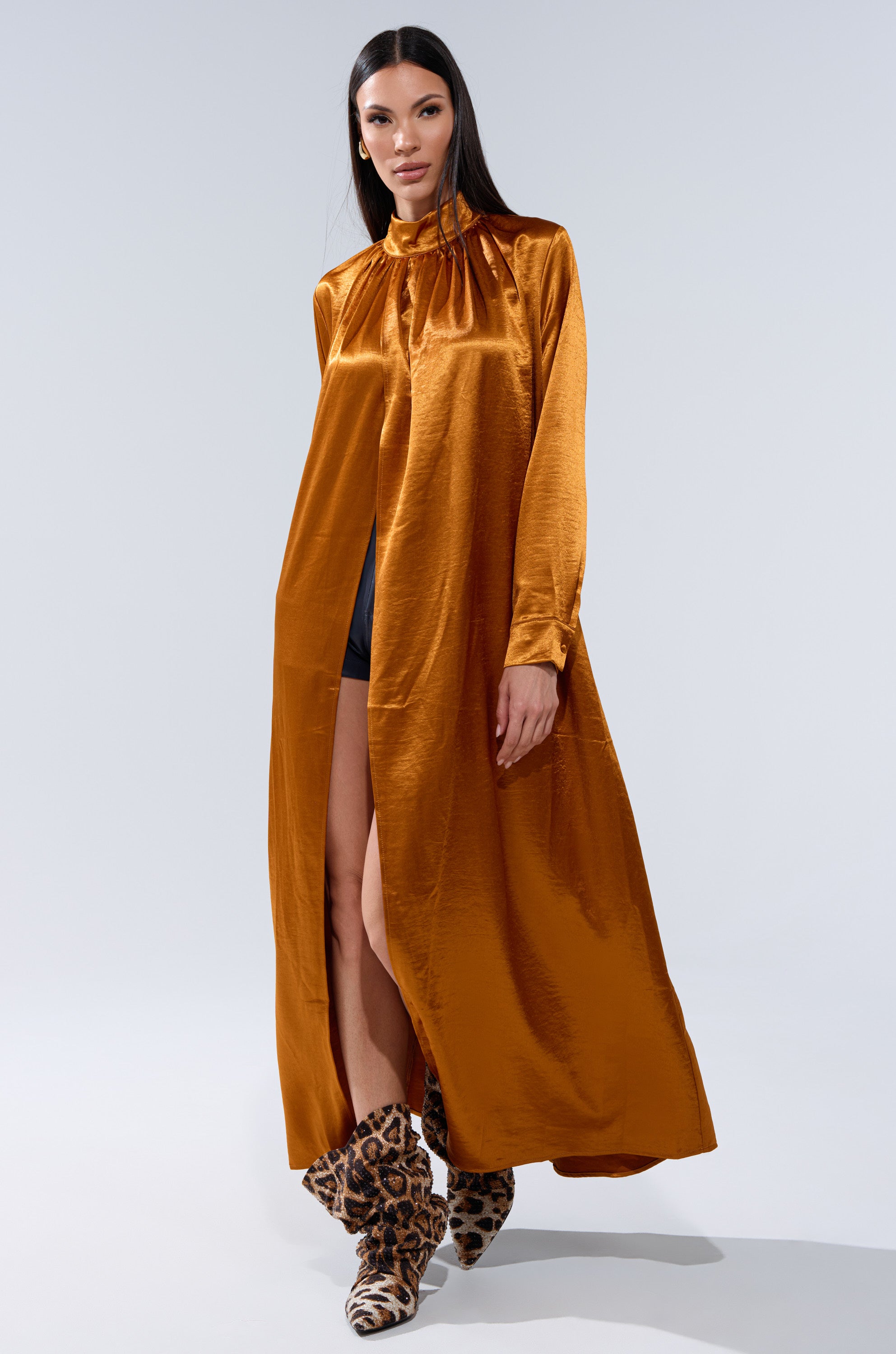 A woman in a long, shiny golden-brown QUEEN TYPE BLOUSE with a mock neck and gathered details, paired with leopard-print ankle boots, stands against a plain light background.