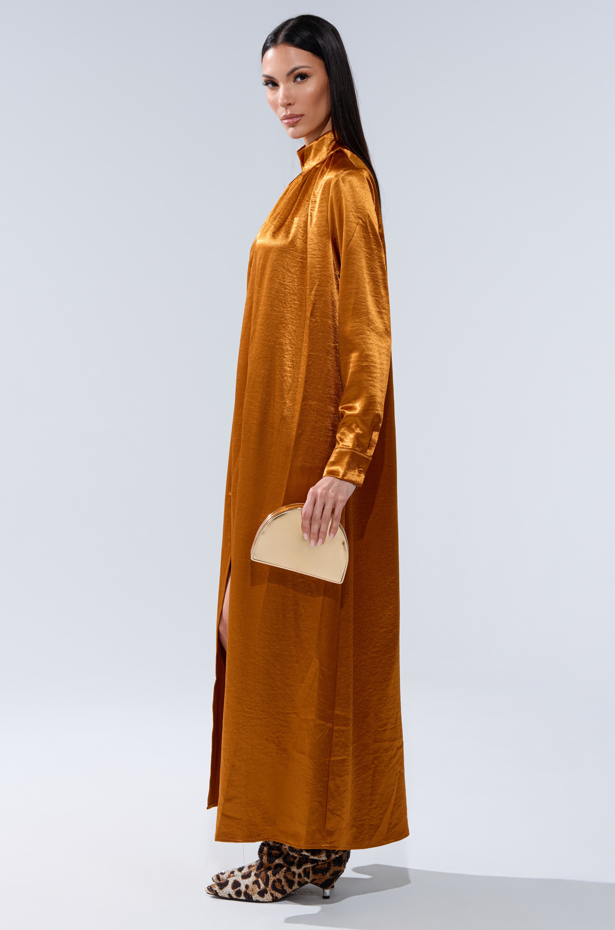 A woman in a long, shiny burnt orange QUEEN TYPE BLOUSE dress with a mock neckline stands in profile, holding a small beige purse. She wears leopard-print ankle boots and has straight dark hair against a plain light background.