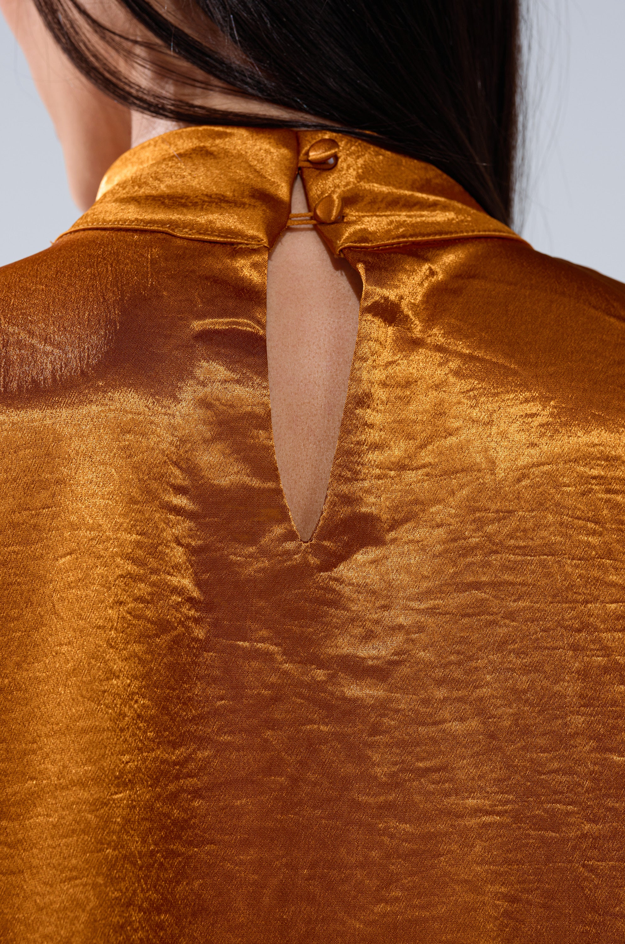 Close-up of the back of a person wearing the QUEEN TYPE BLOUSE, a shiny golden-brown satin top with a high neck, teardrop keyhole cutout, and single button closure at the collar.
