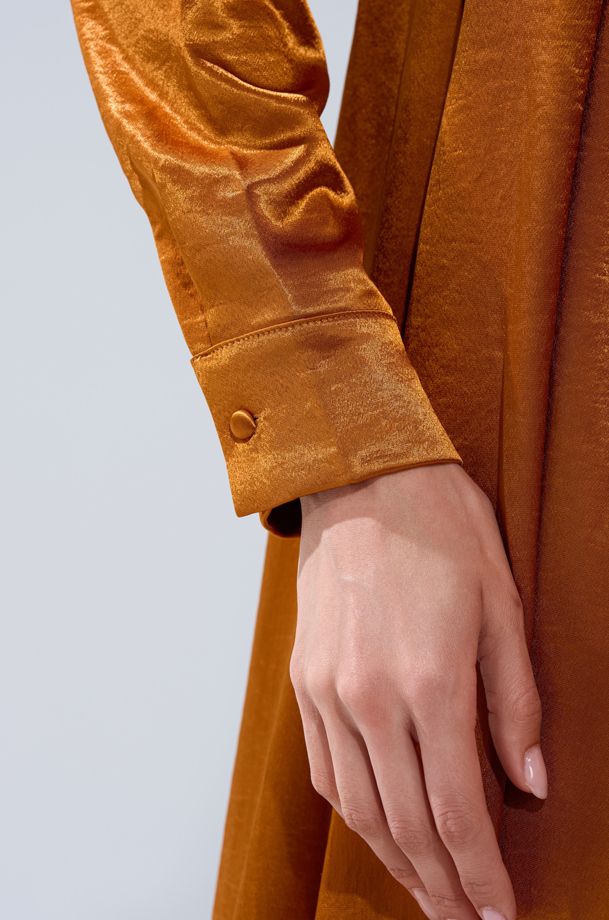A hand rests on the QUEEN TYPE BLOUSE, an orange, shiny satin long sleeve top with silky textured fabric and a cuff finished with a small round button.