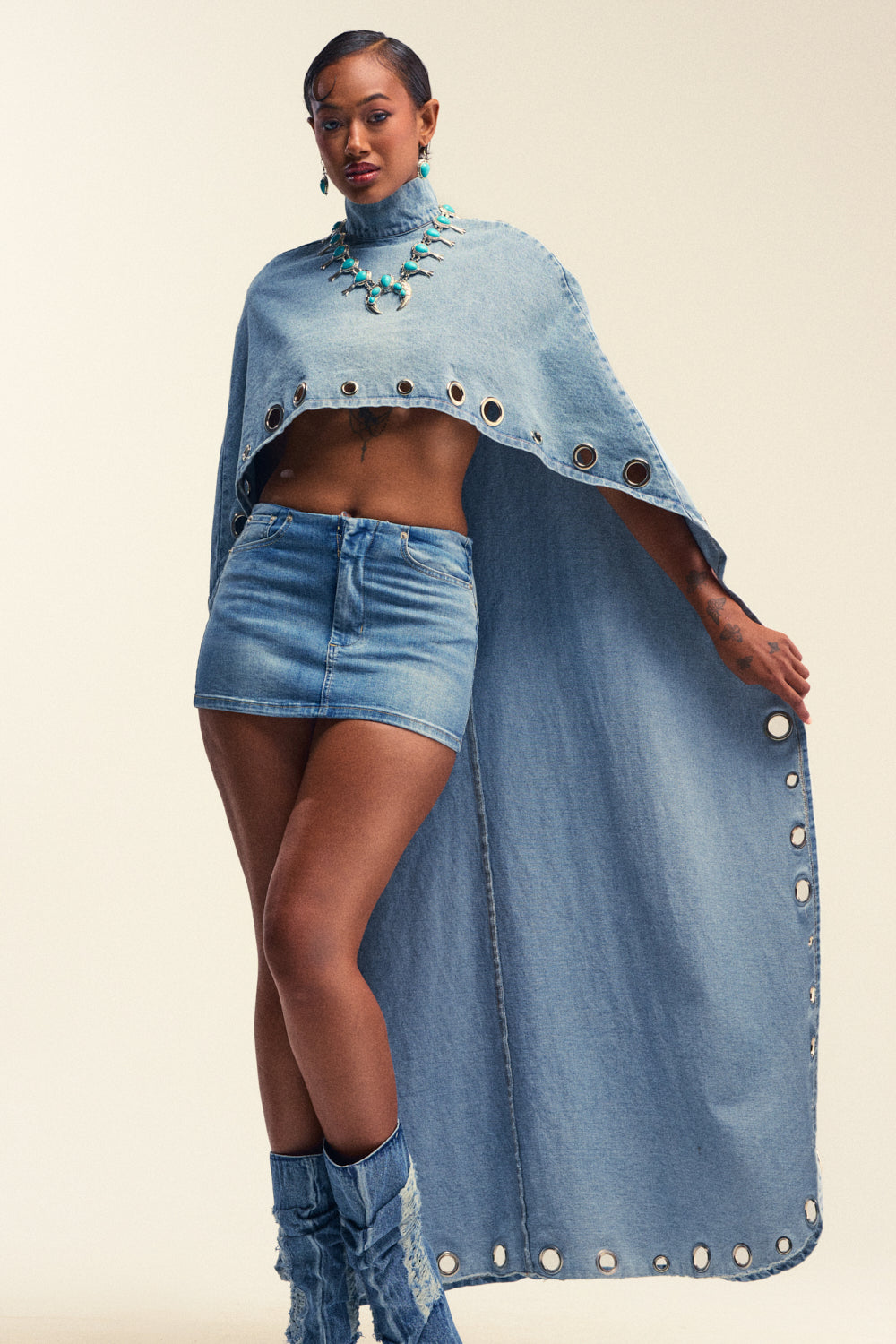 A woman wears the IN YOUR DREAMS DENIM CAPE WITH GROMMETS, paired with a denim mini skirt, denim boots, turquoise jewelry, and poses confidently against a neutral background.