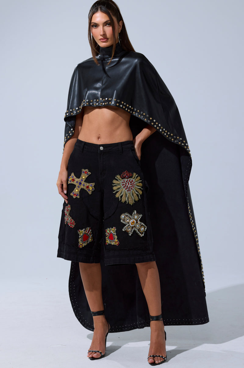 A woman stands confidently in the ALL ABOUT THE DROP TURTLENECK HIGH-LOW BLOUSE, paired with a black leather cape with studded trim, black knee-length shorts featuring large decorative patches, and black high-heeled sandals.
