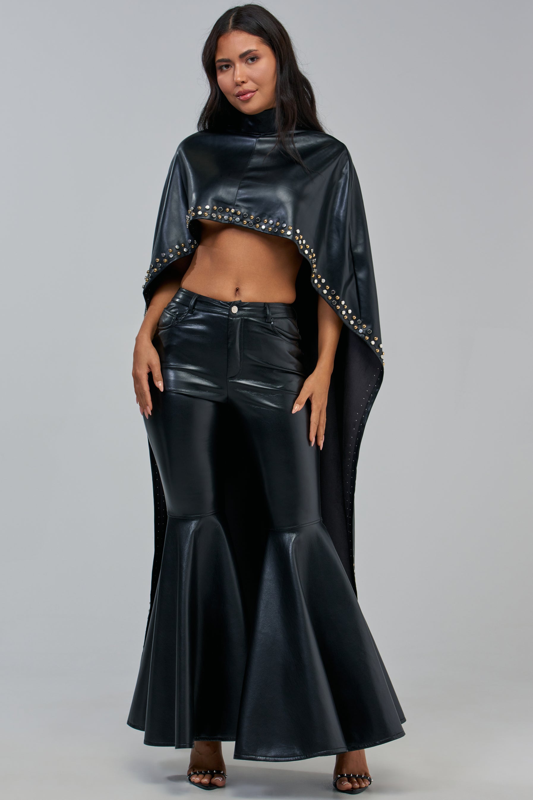 A woman wearing the ALL ABOUT THE DROP TURTLENECK HIGH-LOW BLOUSE styled as a shiny, black high-low cape over a matching high-waisted flared skirt and sandals, standing against a plain light gray background.