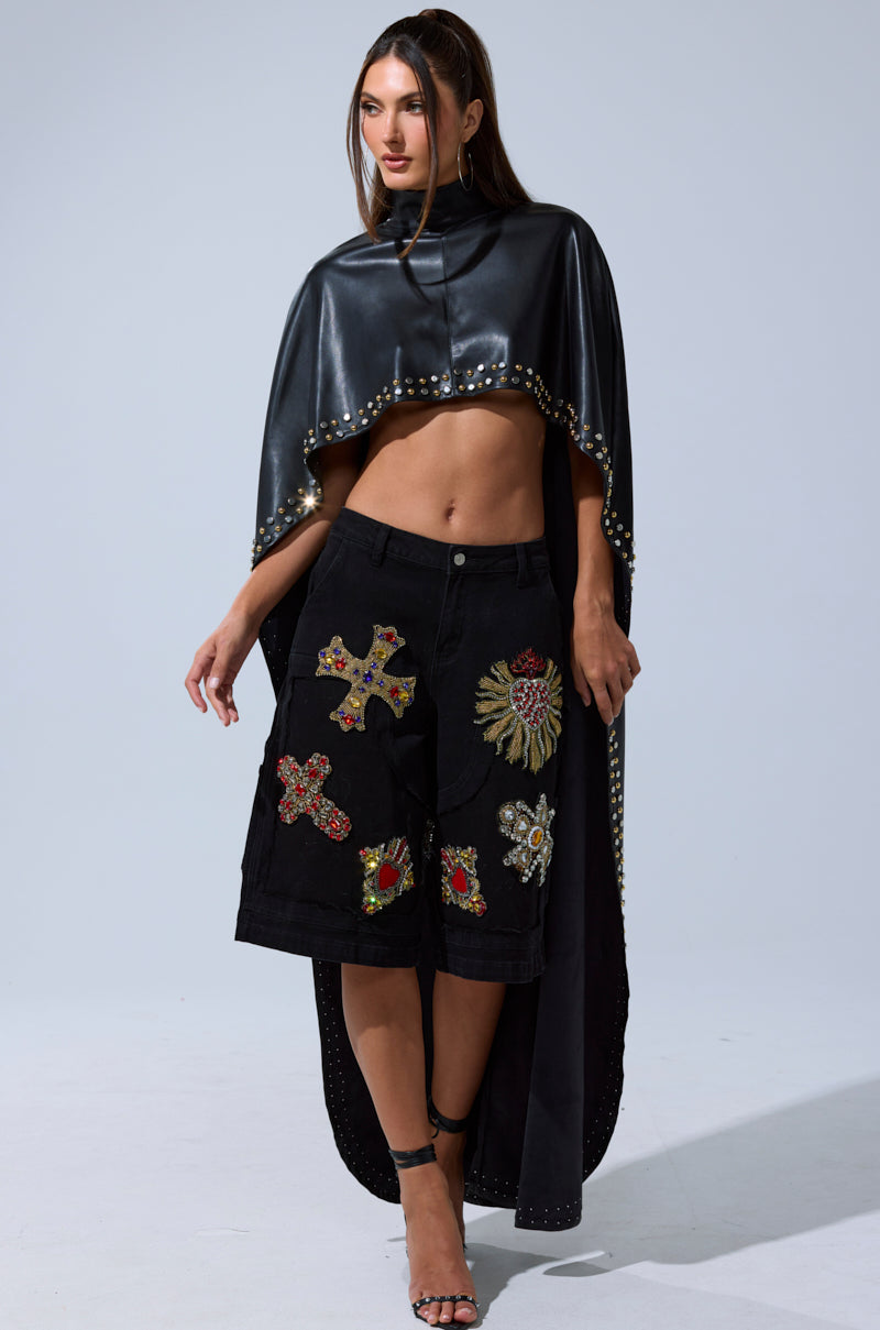 A woman models the ALL ABOUT THE DROP TURTLENECK HIGH-LOW BLOUSE, a black, shiny faux leather cropped cape with silver studs on the shoulders, paired with black knee-length shorts with colorful patches and black strappy heels.