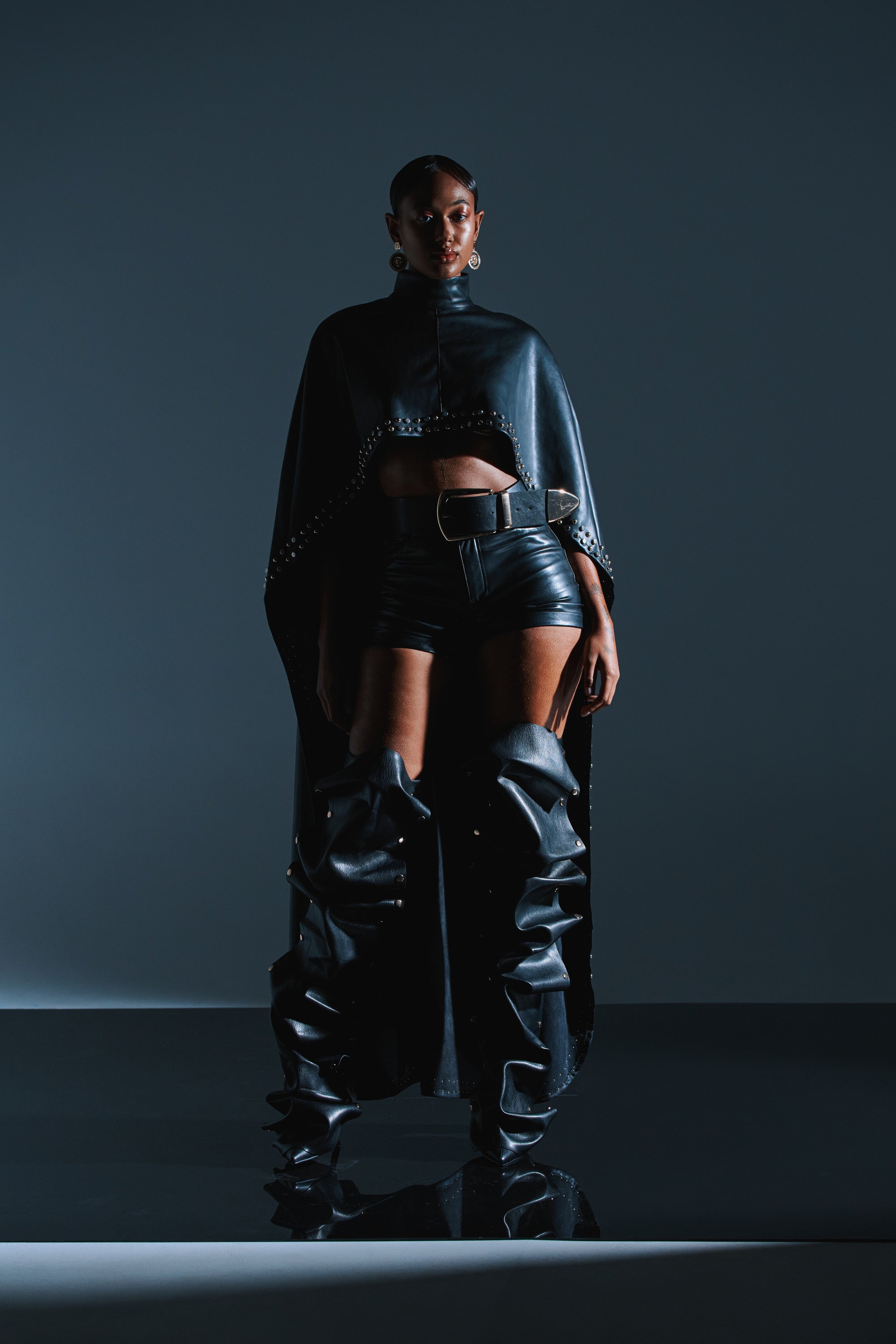 A woman stands confidently in a shadowy studio, wearing the ALL ABOUT THE DROP TURTLENECK HIGH-LOW BLOUSE paired with high-waisted shorts and tall thigh-high boots, her bold look dramatically highlighted by moody lighting.