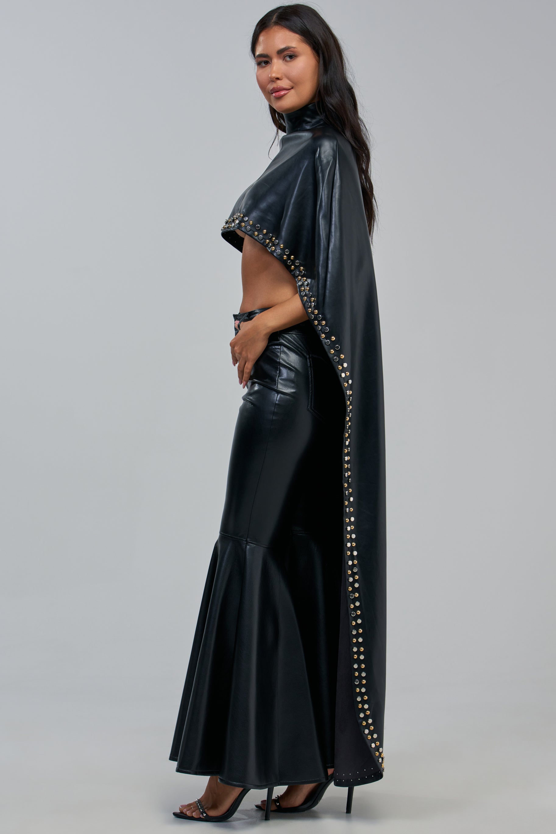 A woman stands sideways against a plain light gray background, wearing the ALL ABOUT THE DROP TURTLENECK HIGH-LOW BLOUSE—a black cape-style blouse adorned with gold studs—paired with a matching floor-length skirt featuring a flared hem and black heels.