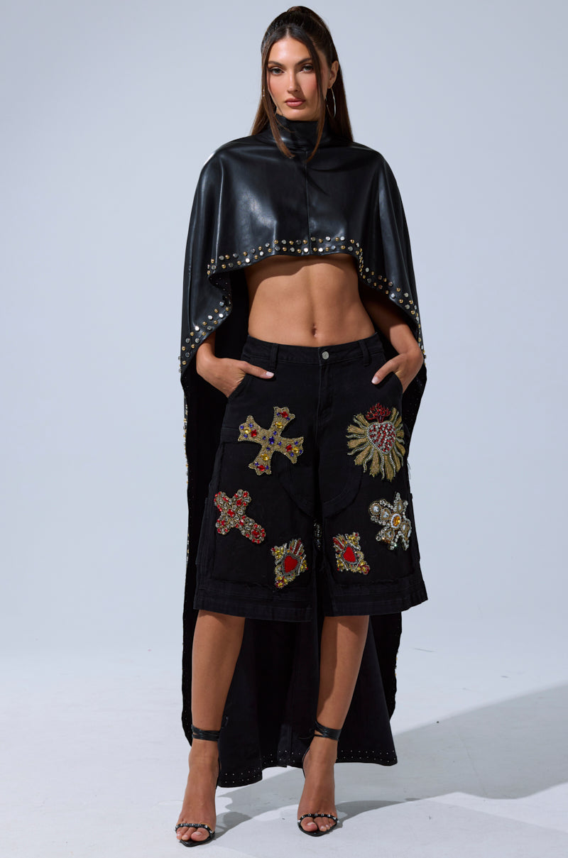 A woman stands confidently in the ALL ABOUT THE DROP TURTLENECK HIGH-LOW BLOUSE, styled as a cape, paired with a knee-length black skirt featuring colorful embroidered patches and high-heeled sandals against a plain light background.