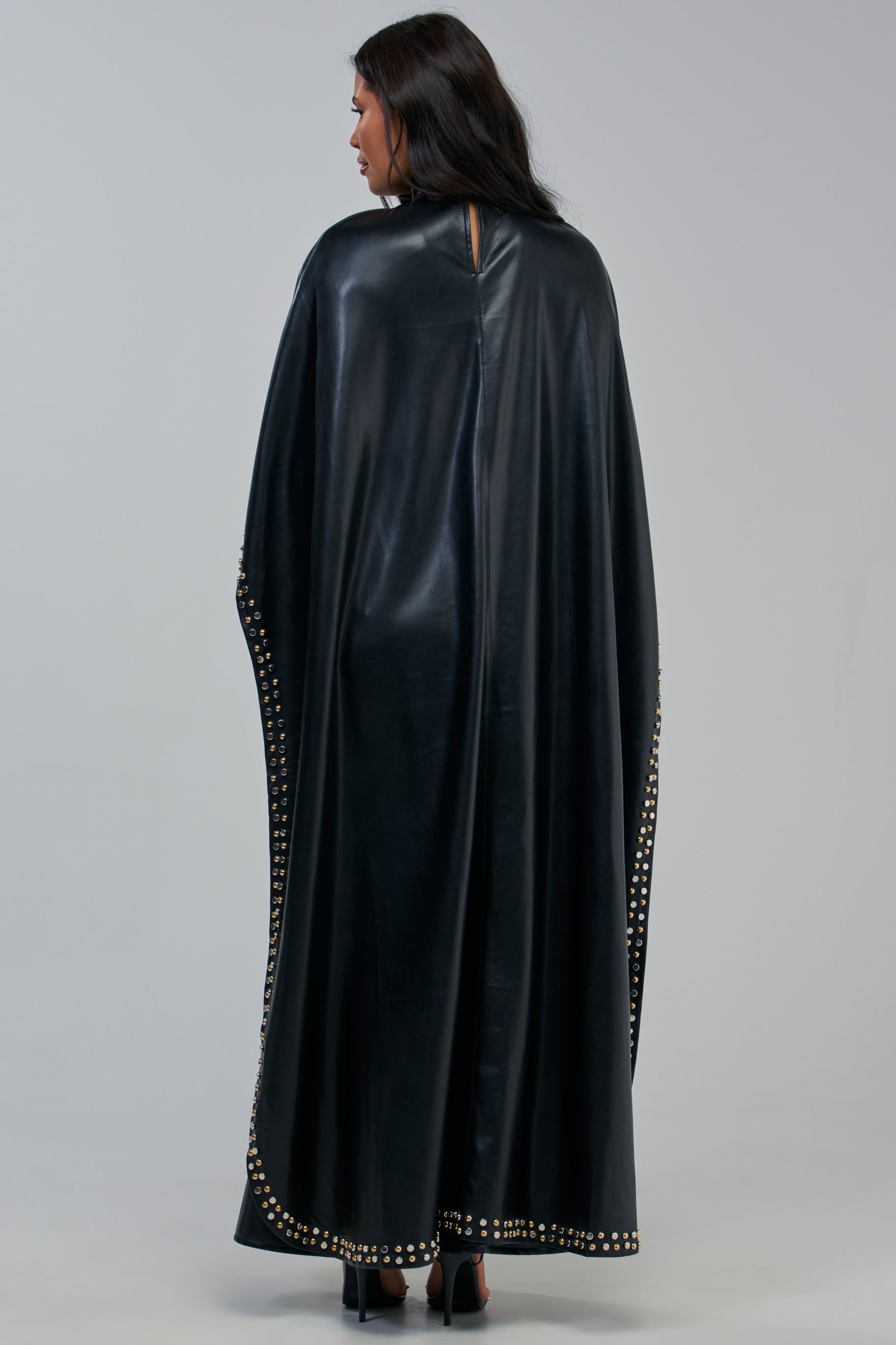A person stands facing away, wearing the ALL ABOUT THE DROP TURTLENECK HIGH-LOW BLOUSE in black, which features gold studs accenting the edges. They pair it with black high heels against a plain light gray background.