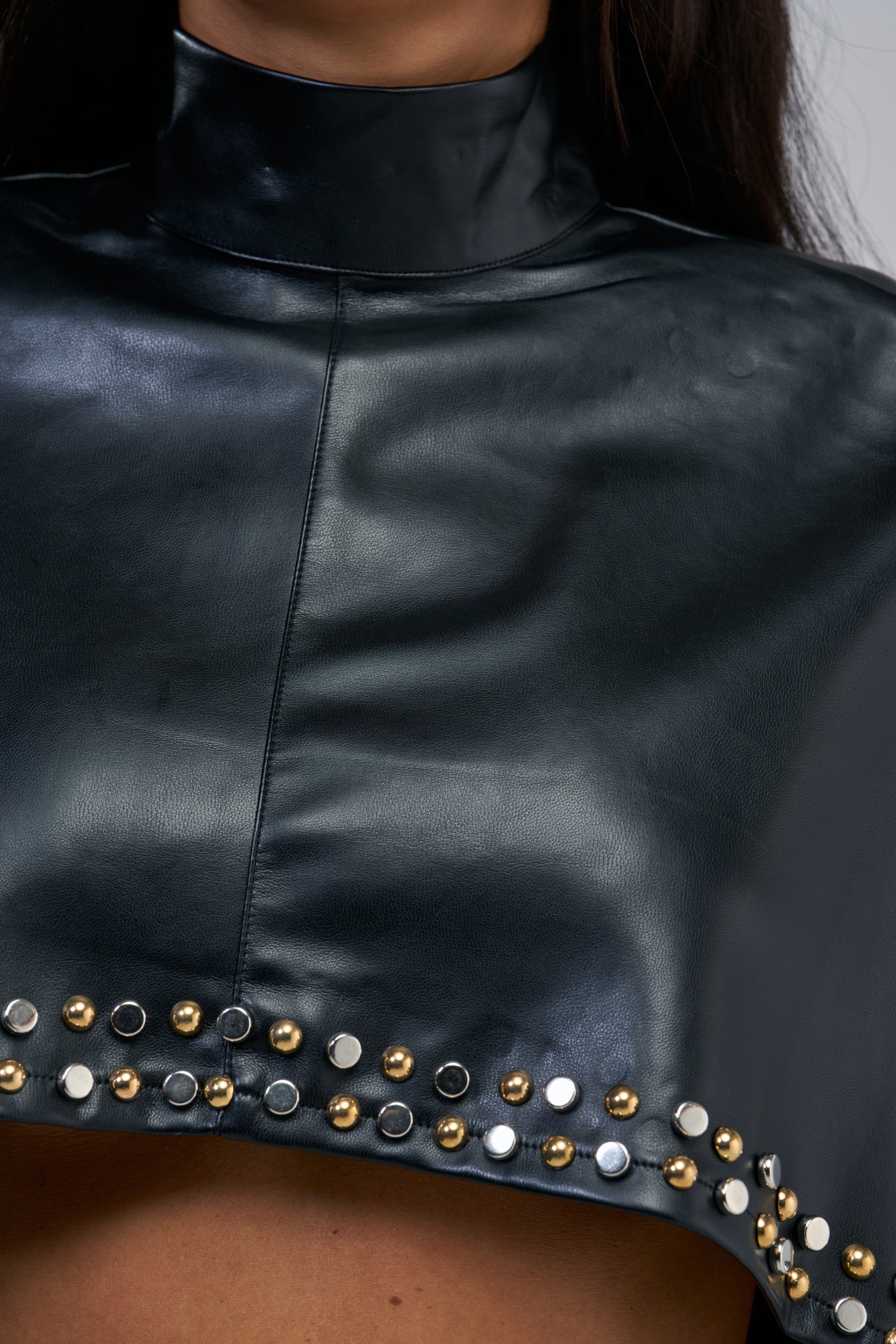 A close-up of a person wearing the ALL ABOUT THE DROP TURTLENECK HIGH-LOW BLOUSE—a black faux leather blouse with a high turtleneck collar and a dramatic hem adorned with gold, silver, and black round studs in a striking row, showcasing the bold style of this studded cape-inspired blouse.
