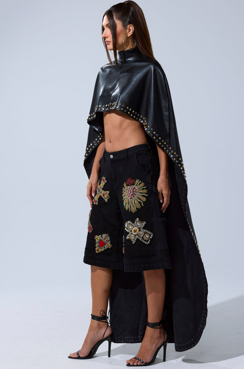 A woman poses confidently against a plain background in the ALL ABOUT THE DROP TURTLENECK HIGH-LOW BLOUSE, styled over her bare torso with embroidered black knee-length shorts and strappy heels.