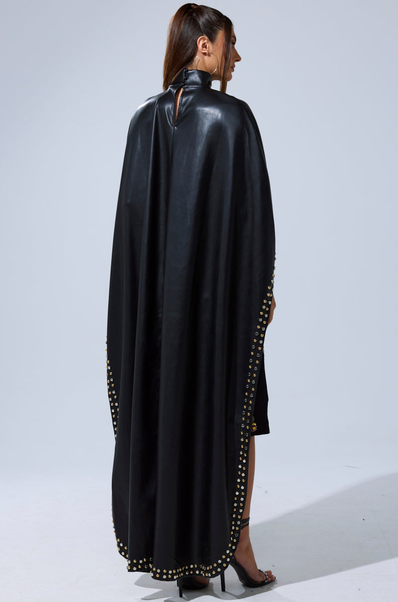 A woman with a ponytail models the ALL ABOUT THE DROP TURTLENECK HIGH-LOW BLOUSE, featuring gold studded edges. She pairs it with a black dress and heels against a plain light gray background.