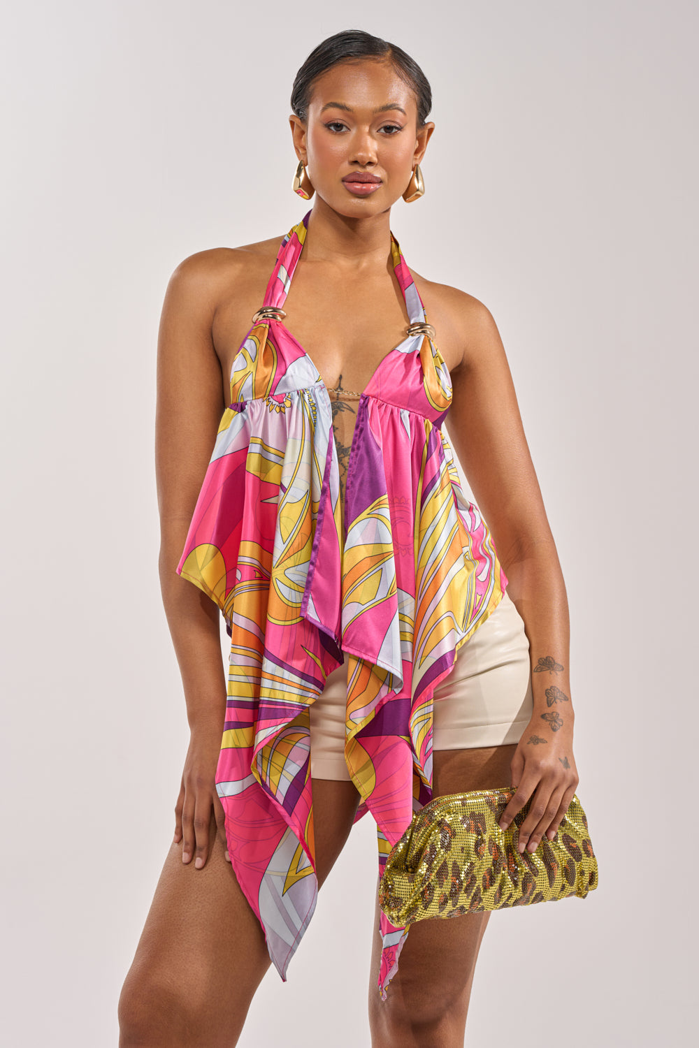 A woman poses confidently in the FUN AND FLIRTY SATIN BLOUSE with pink, yellow, and white patterns, paired with cream shorts, gold hoop earrings, and holding a green and gold leopard-print clutch against a plain light background. 1