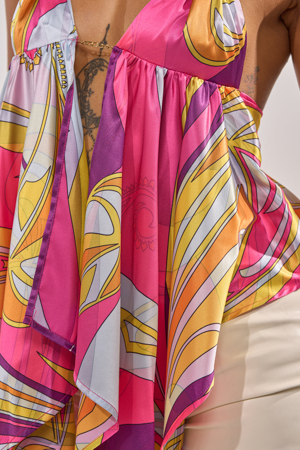 A close-up of someone wearing the FUN AND FLIRTY SATIN BLOUSE—a colorful, patterned satin halter top in pink, yellow, orange, and white with draped fabric and a peek of chest tattoo—ideal for summer nightlife looks. 7