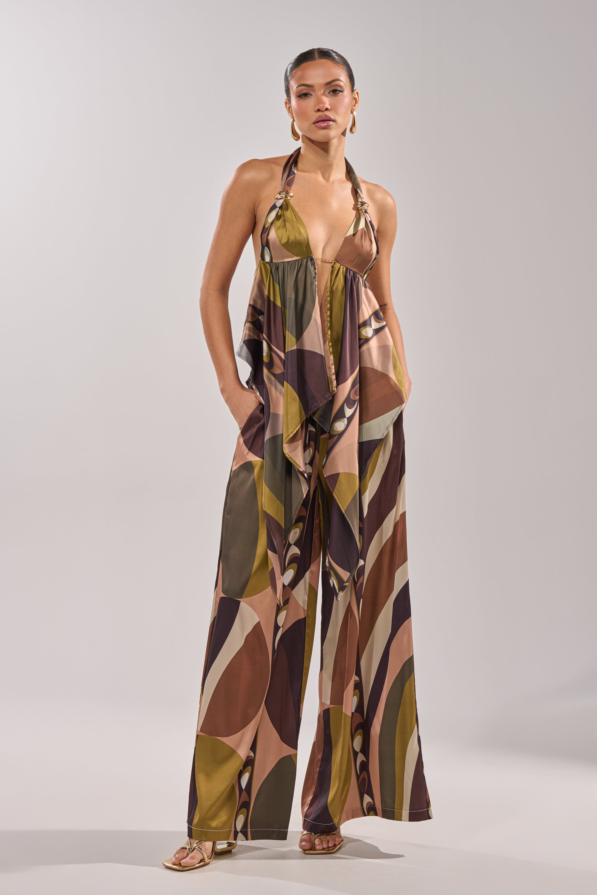 A woman stands confidently in the WILD AND FREE TROUSER, featuring palazzo-style wide legs and an abstract geometric pattern in earthy olive green, brown, and beige. She has her hands in her pockets and wears strappy sandals. 2