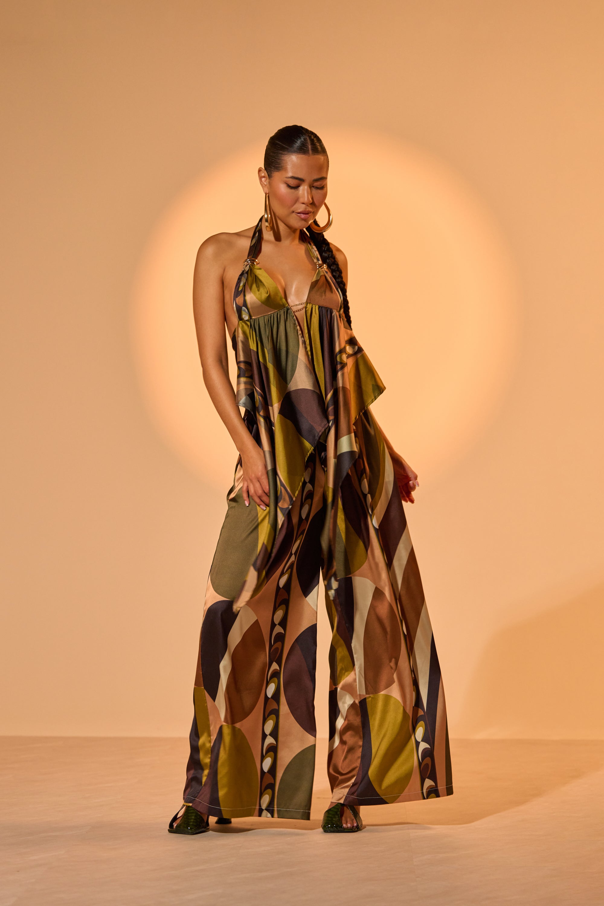 A woman stands confidently before a beige background wearing the WILD AND FREE TROUSER, a palazzo pant with bold tropical print in earthy tones. Her slicked-back hair and large hoop earrings complete the effortlessly chic look. 1