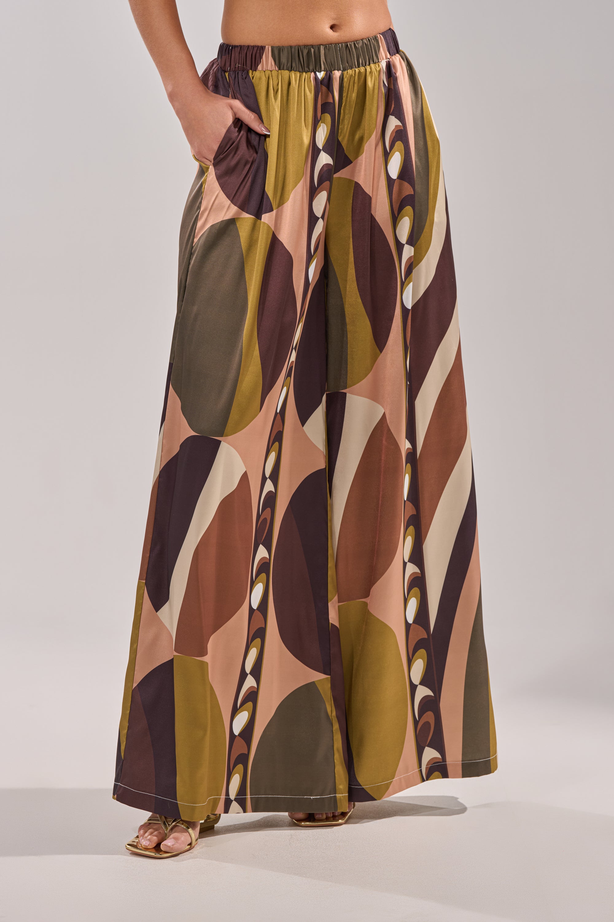 Wearing the WILD AND FREE TROUSER, featuring a high-waisted, wide-leg palazzo cut and an abstract geometric print in earthy tones, a person stands with one hand in the pocket; only their lower body is visible. 3