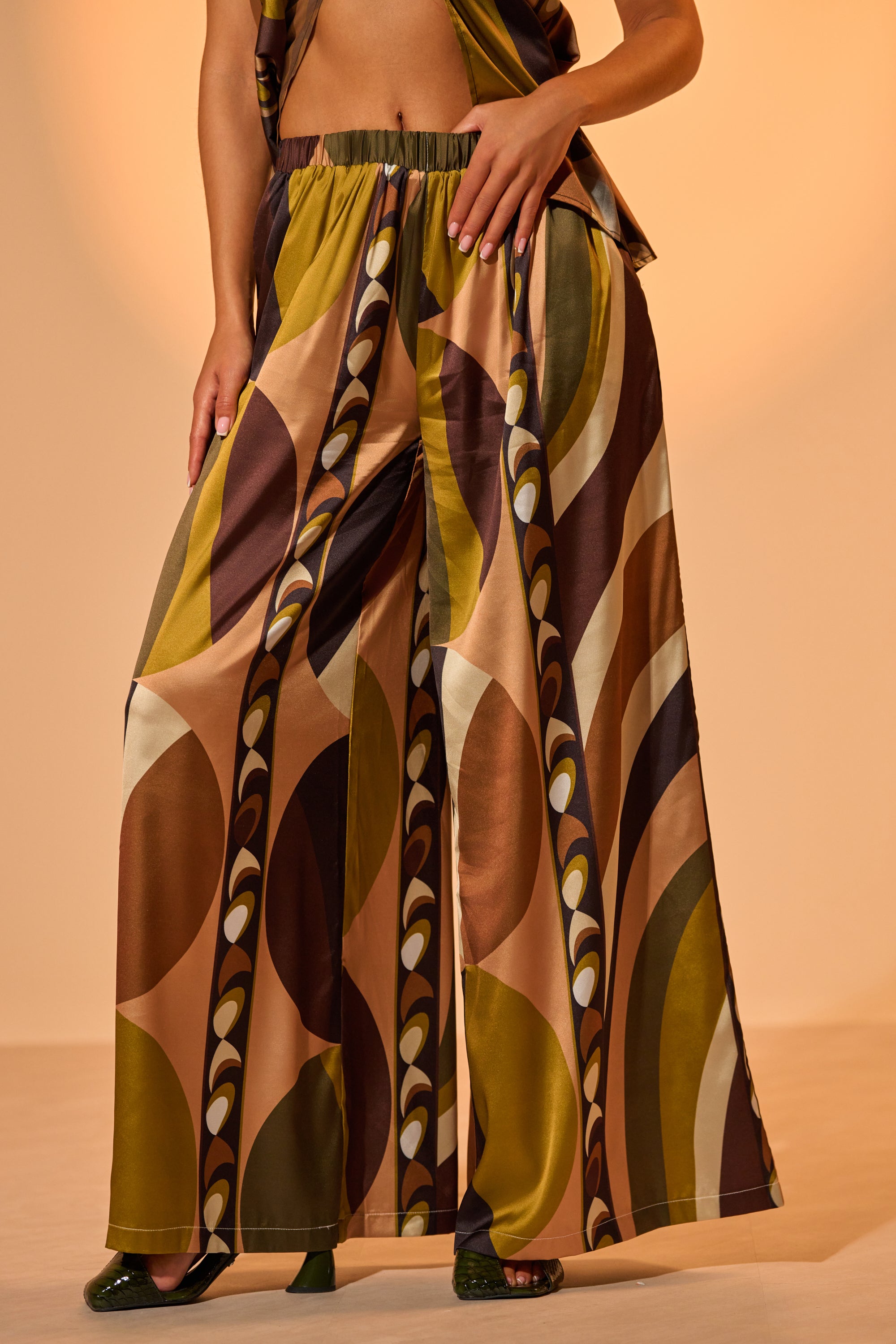 A person stands against a neutral background wearing WILD AND FREE TROUSER—wide-leg palazzo pants featuring a bold, retro geometric pattern in brown, olive green, mustard, and beige. 0