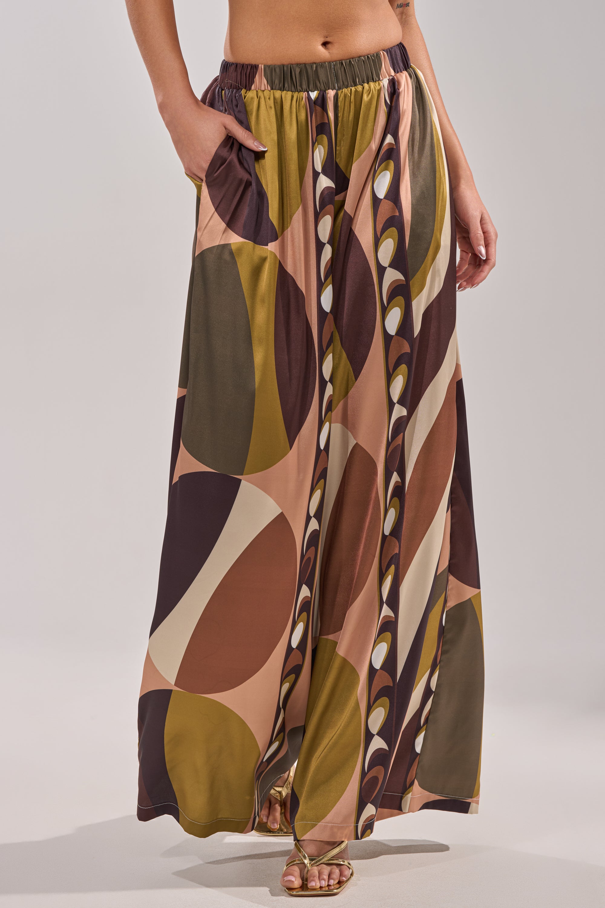 Someone wearing the WILD AND FREE TROUSER, a long, flowy palazzo pant with a bold geometric pattern in brown, olive, pink, and beige. Features an elastic waistband and pockets, styled with sandals for an effortlessly chic look. 4