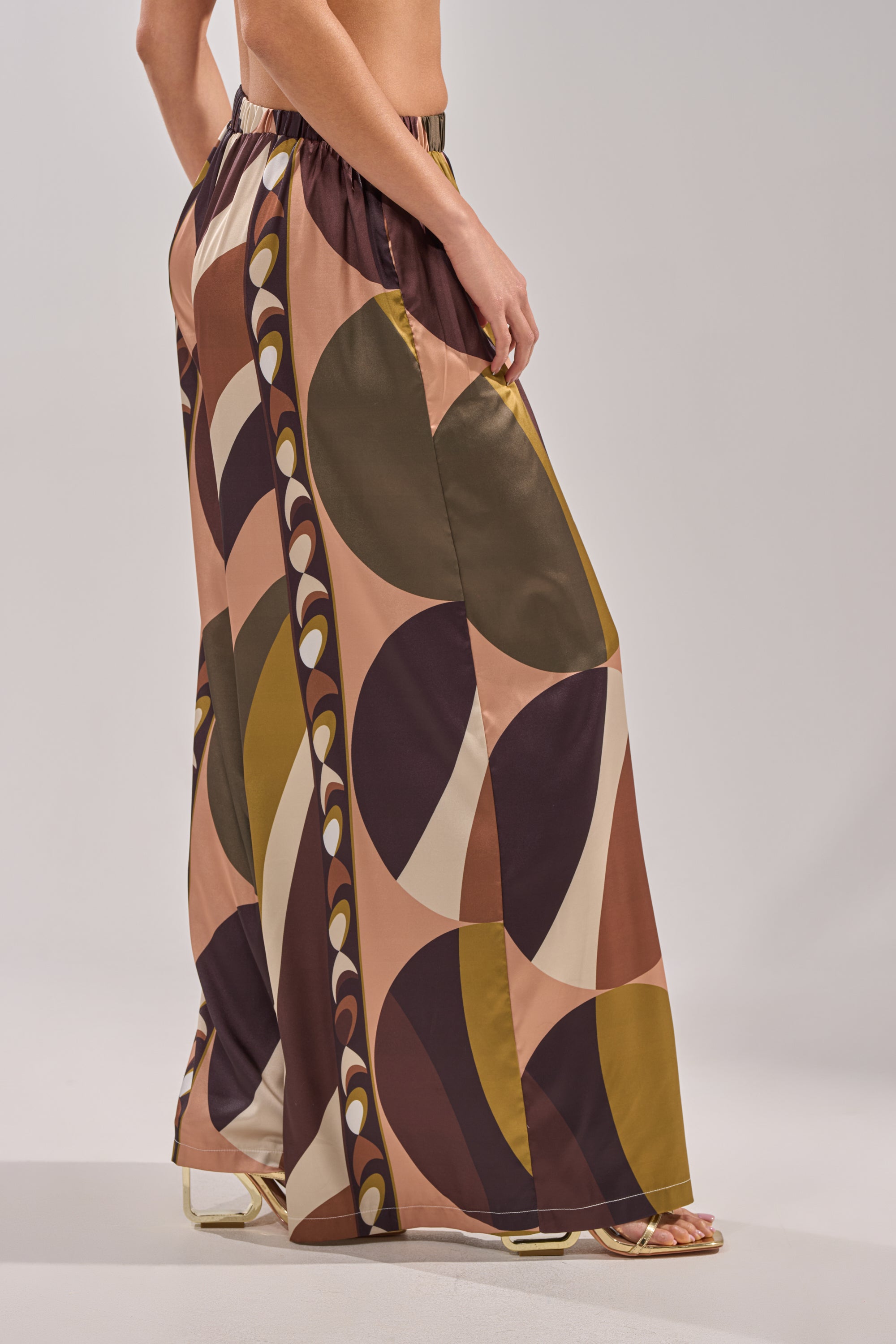 A person in profile stands against a neutral background wearing the WILD AND FREE TROUSER—satin, wide-leg palazzo pants featuring a bold geometric pattern in brown, olive, mustard, and peach. 6