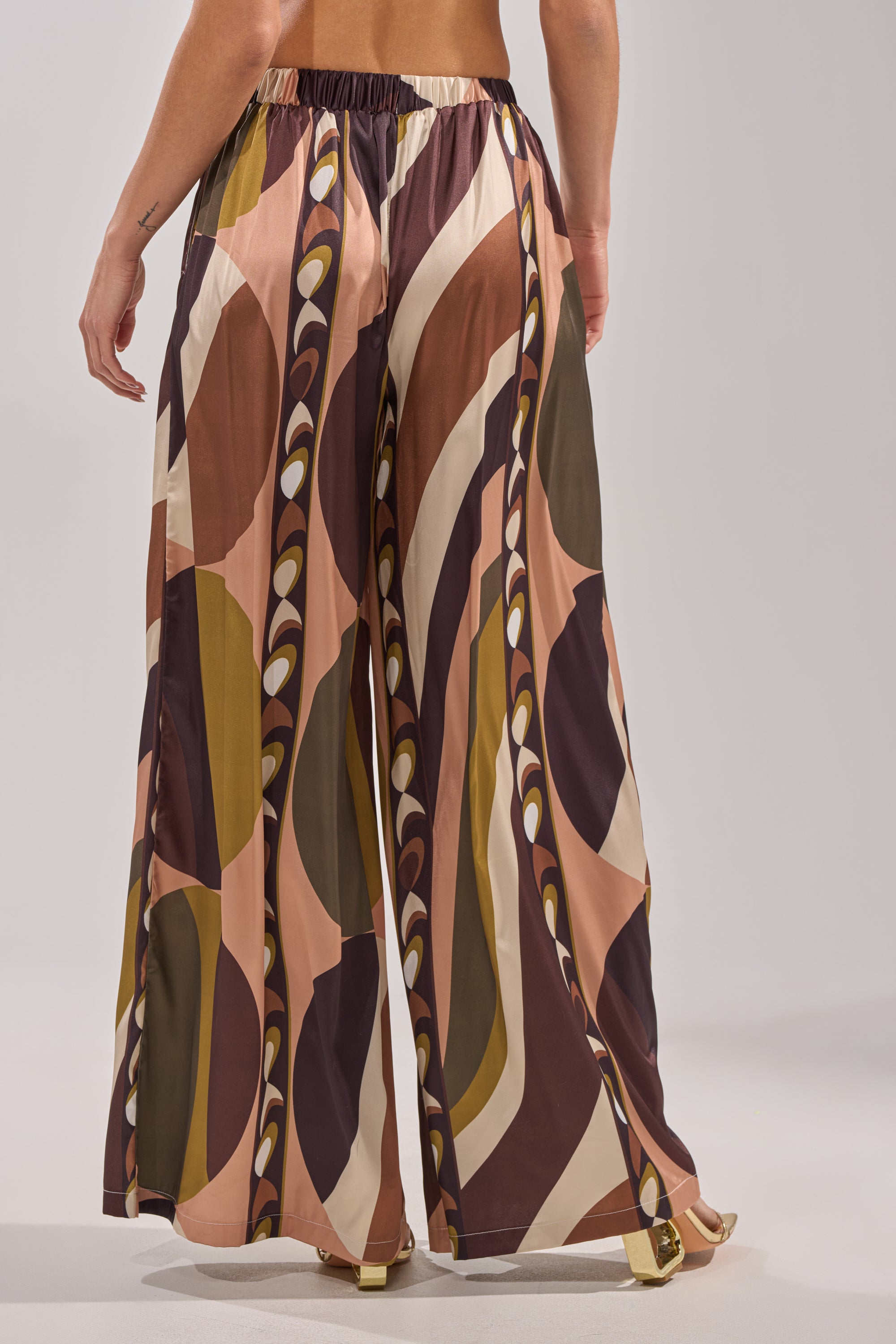 A person models the WILD AND FREE TROUSER—loose, high-waisted palazzo pants featuring a bold, abstract geometric print in brown, pink, olive, and cream. The photo is waist-down from the back. 7