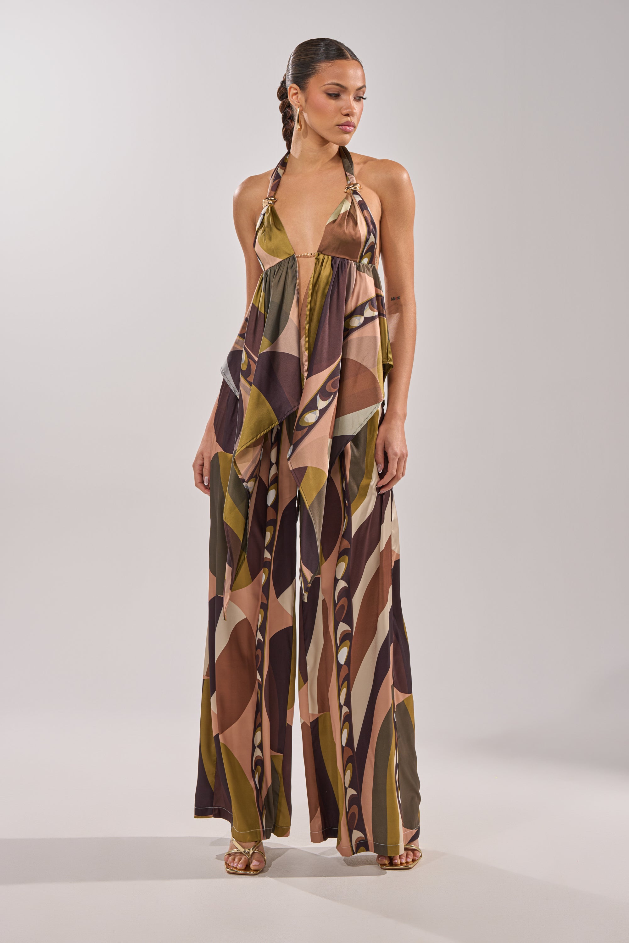 A woman wears the WILD AND FREE TROUSER, a flowing halter-neck satin jumpsuit with palazzo pants and a geometric pattern in brown, olive, and beige, standing against a plain light gray background. 8
