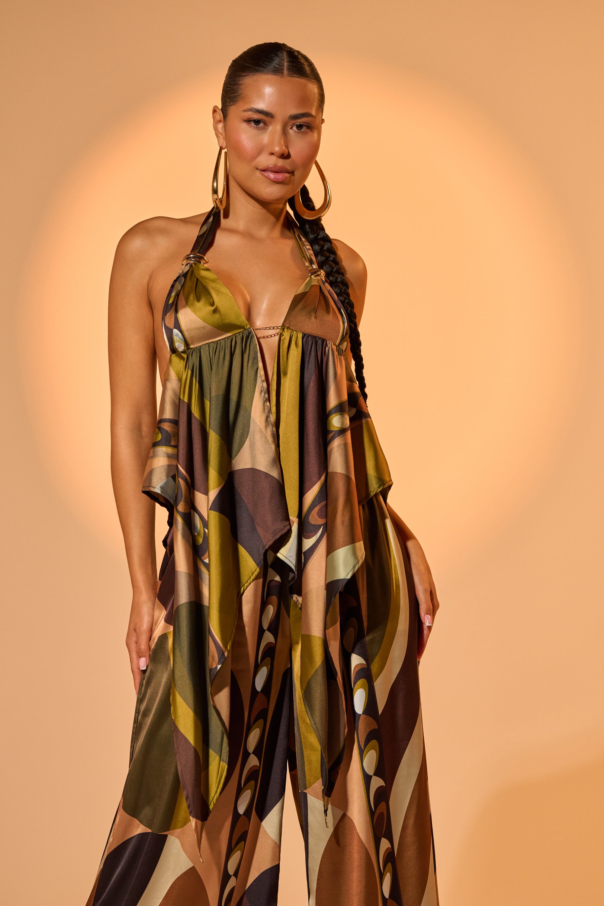 A woman with long braided hair stands confidently in front of a softly lit background, wearing the WILD AND FREE BLOUSE with an abstract print and accessorized with large hoop earrings.