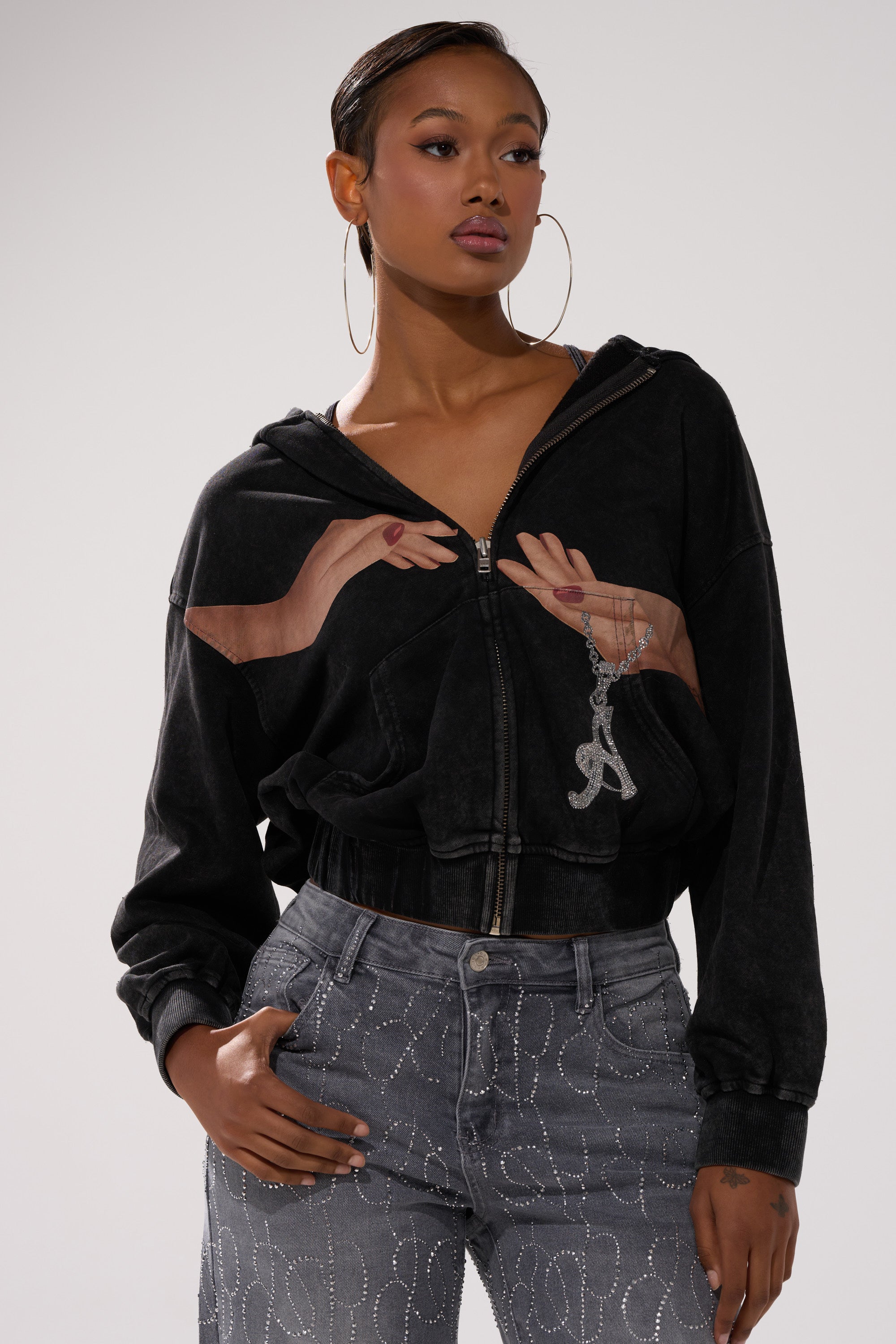 A woman wearing hoop earrings, gray swirl-pattern jeans, and the GRAB MY HAND ZIP UP SWEATSHIRT with a hands-holding-pendant graphic poses against a plain light background. 0