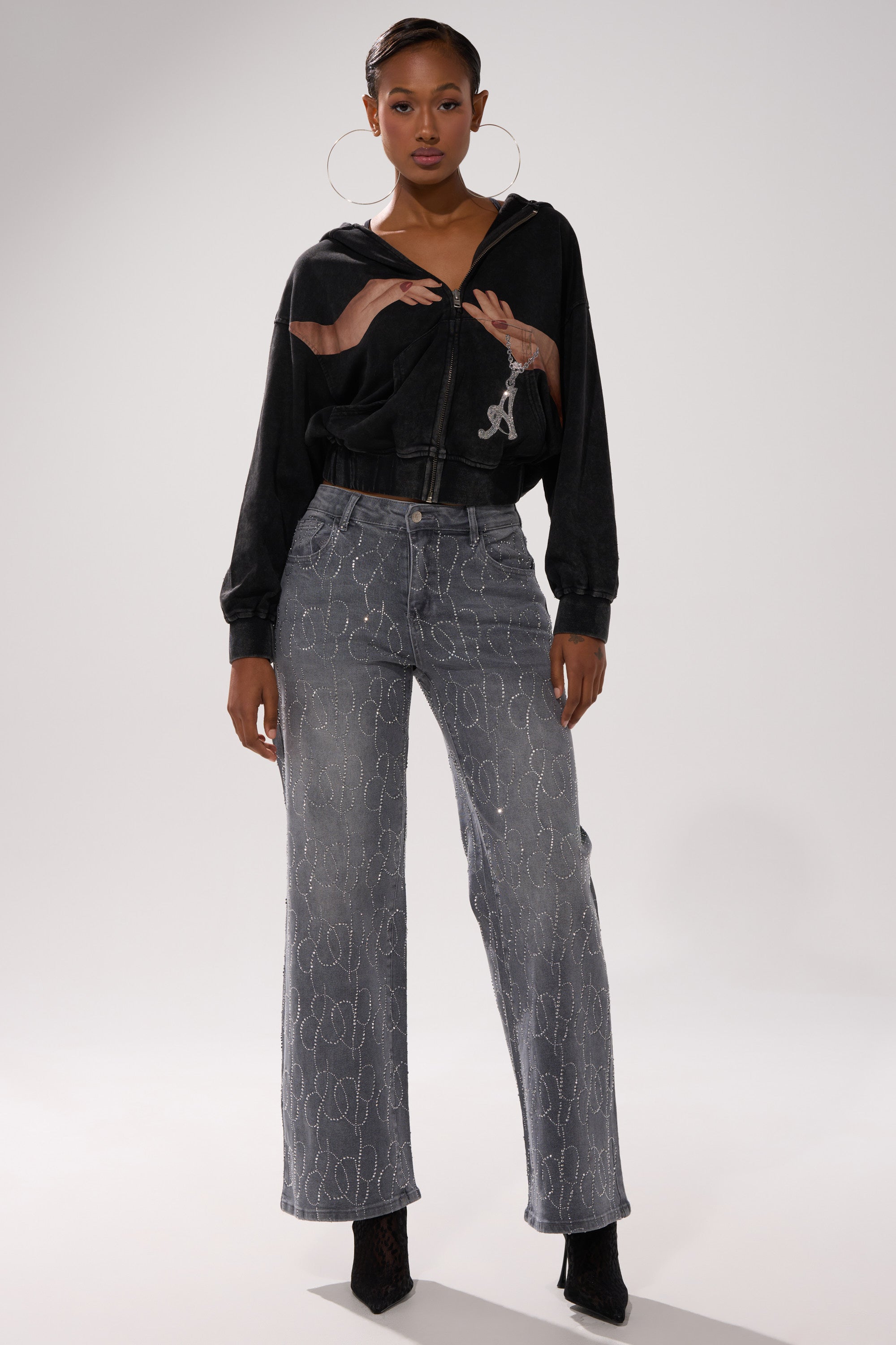 A woman poses confidently in the black GRAB MY HAND ZIP UP SWEATSHIRT with a hand graphic, paired with gray high-waisted patterned jeans, large hoop earrings, and black heeled boots against a plain light background. 6