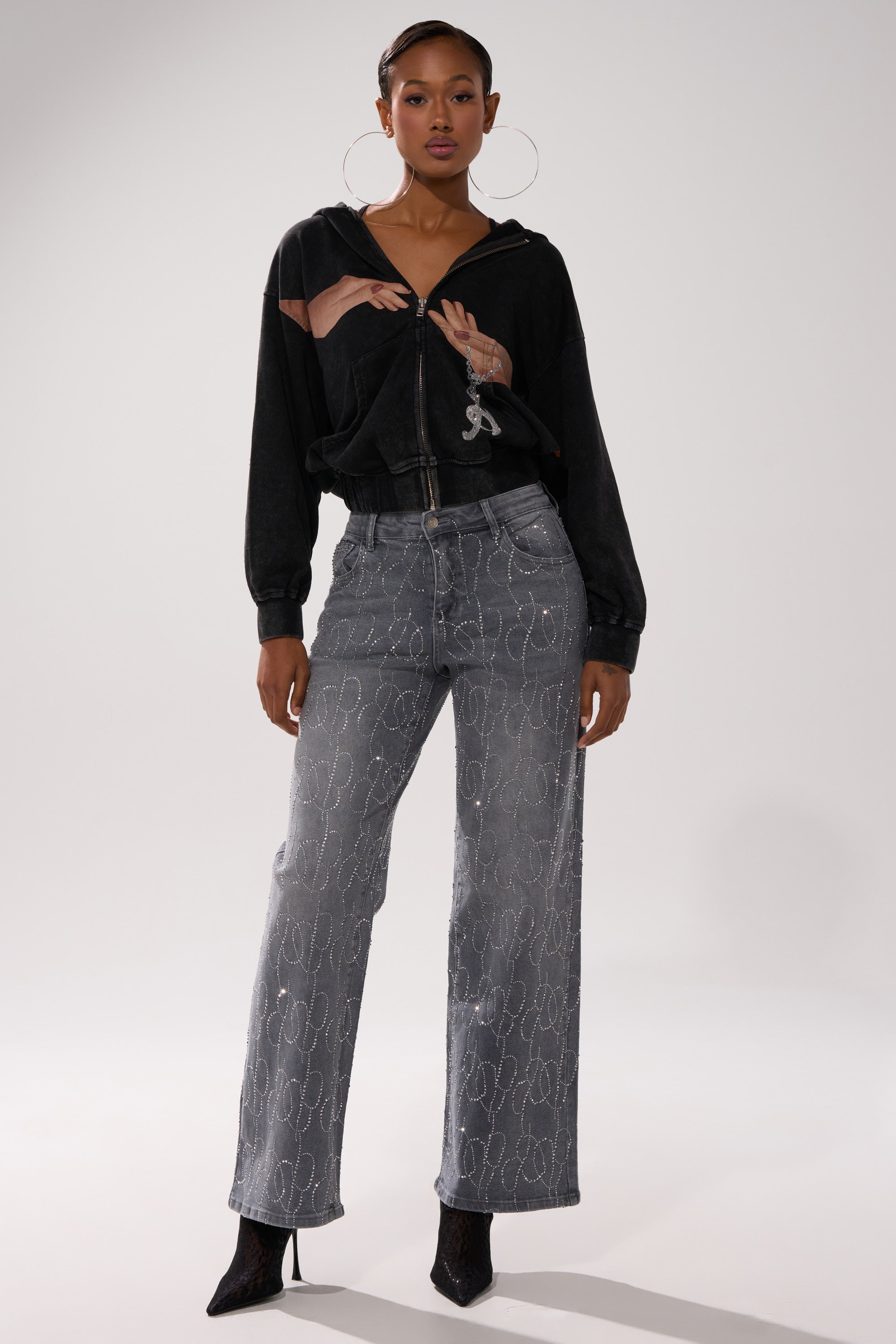 A woman stands confidently in the GRAB MY HAND ZIP UP SWEATSHIRT, paired with light blue high-waisted jeans with a chain-link pattern, black heeled boots, and large hoop earrings. 7