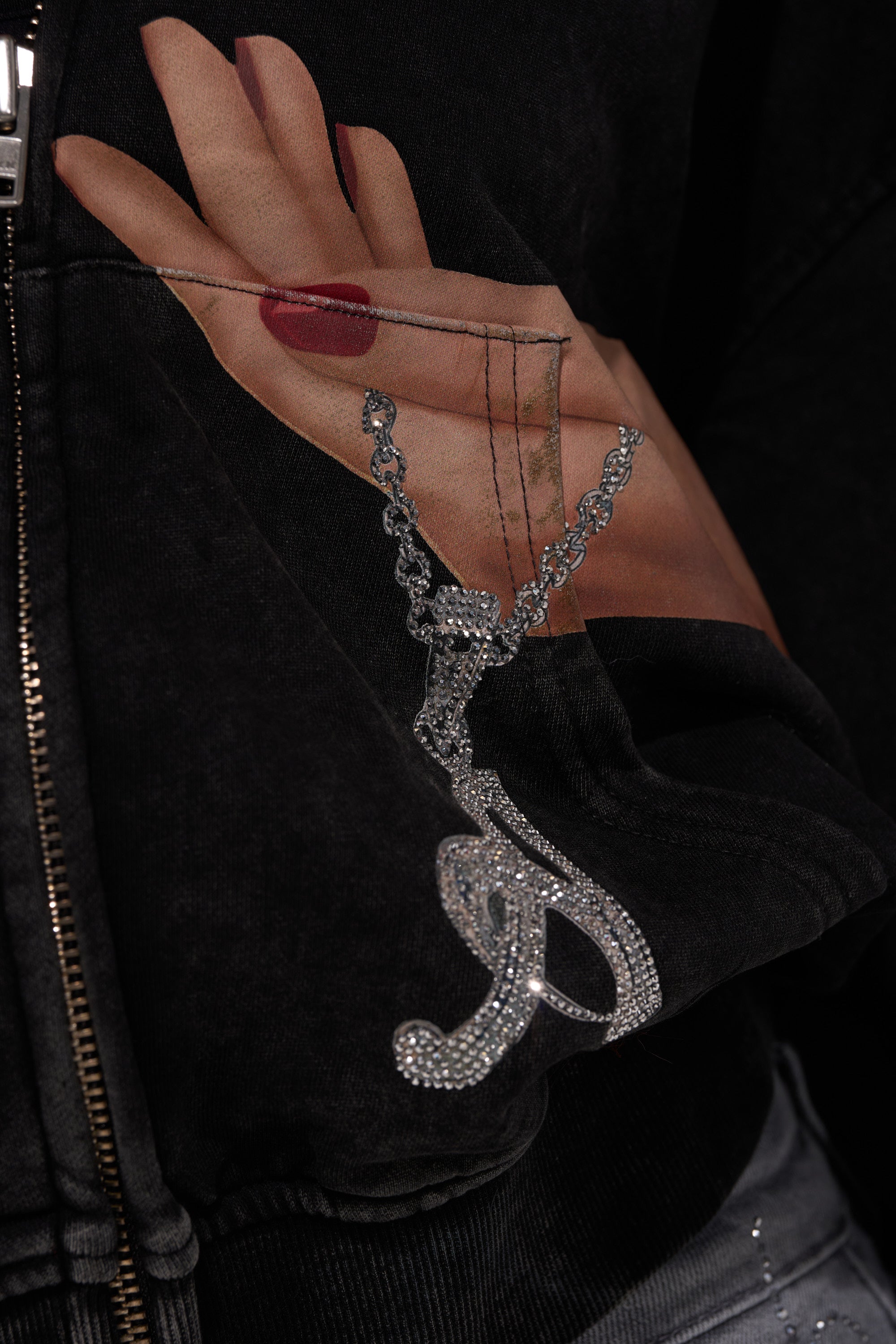 A close-up of the AKIRA Label GRAB MY HAND ZIP UP SWEATSHIRT, showcasing a graphic hand with red nails holding a sparkling silver chain and pendant, draped realistically over the black fabric. 8