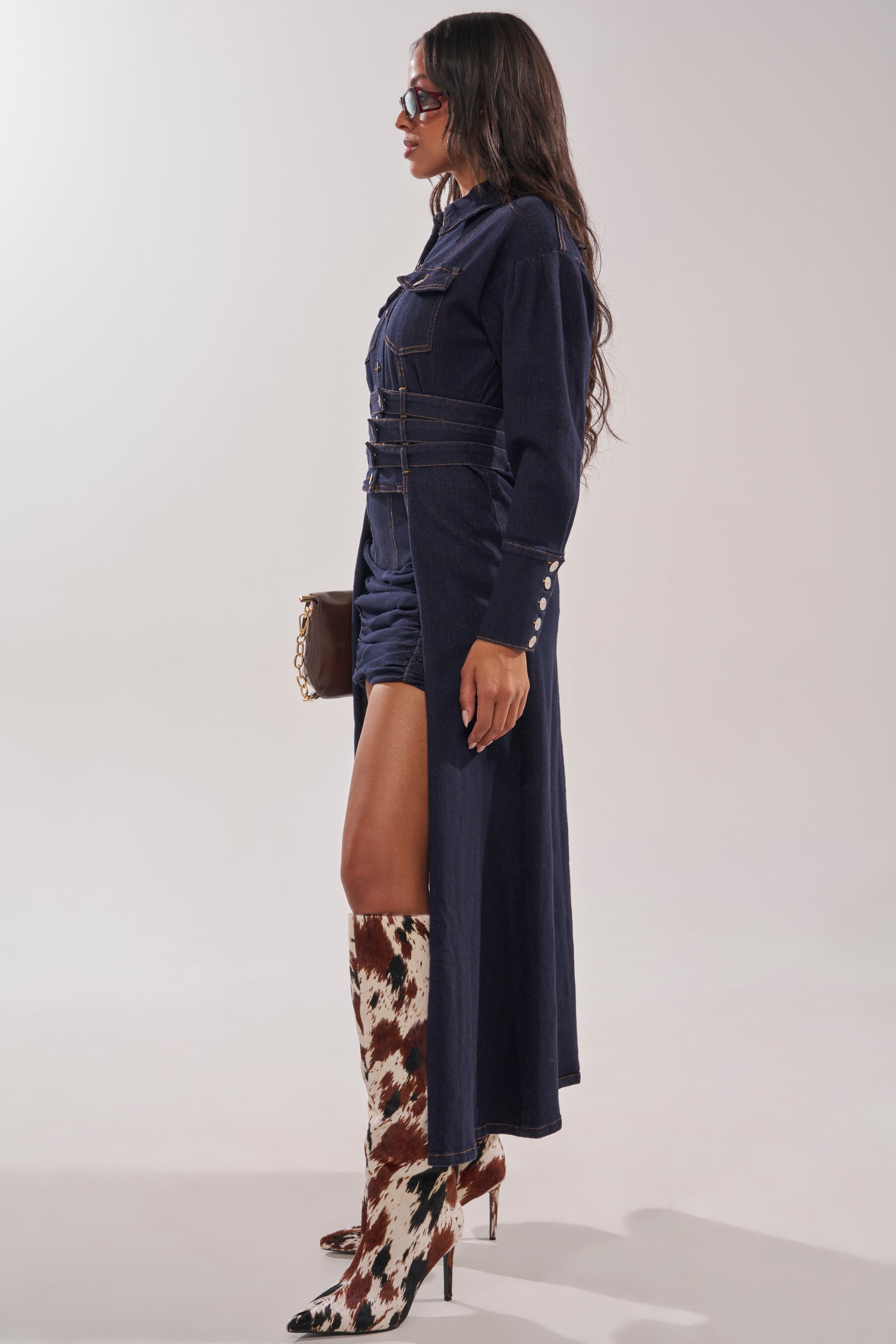 Woman wearing the CITY NIGHTS DENIM BUTTON DOWN styled as a dress with a high side slit, paired with patterned knee-high boots, sunglasses, and a small brown bag, standing against a plain background. 3