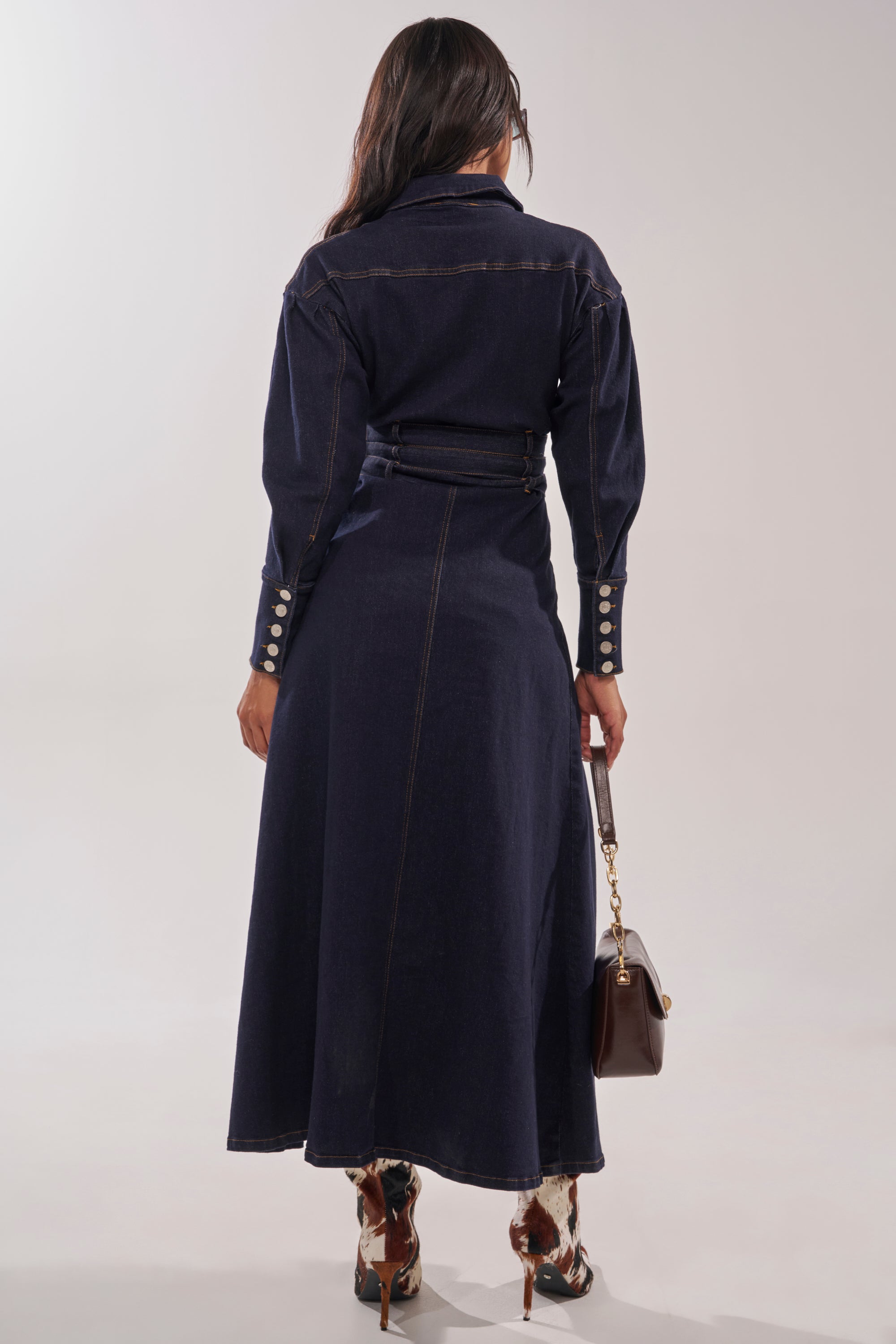 A woman faces away, wearing the CITY NIGHTS DENIM BUTTON DOWN as a dark longline dress with visible seams and buttoned cuffs. She holds a brown handbag and pairs it with heeled boots in a bold brown and white pattern. 4
