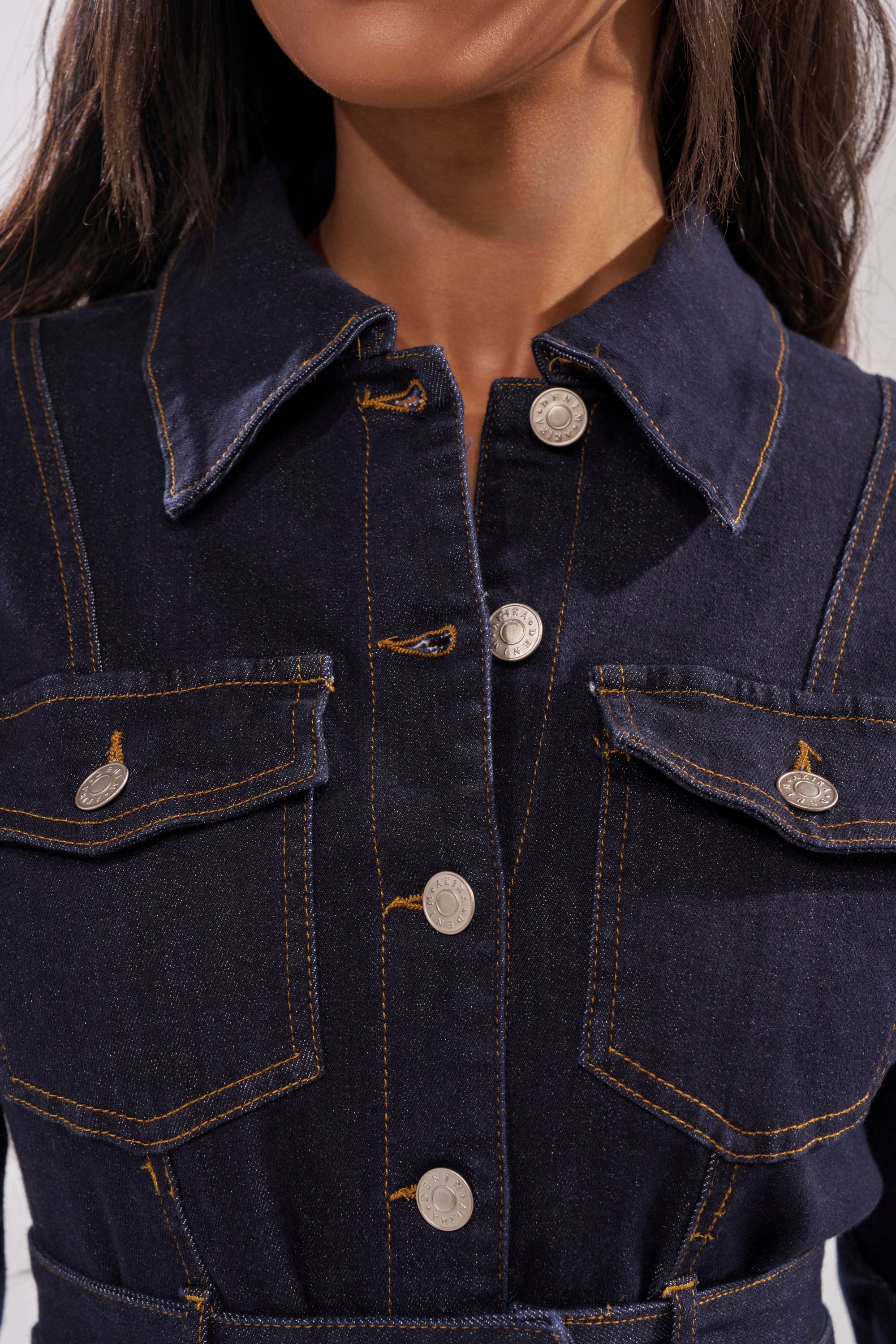 A close-up of a woman wearing the CITY NIGHTS DENIM BUTTON DOWN, featuring dark-wash denim, tan stitching, silver buttons, and two buttoned chest pockets. Her face is partially out of frame. 5