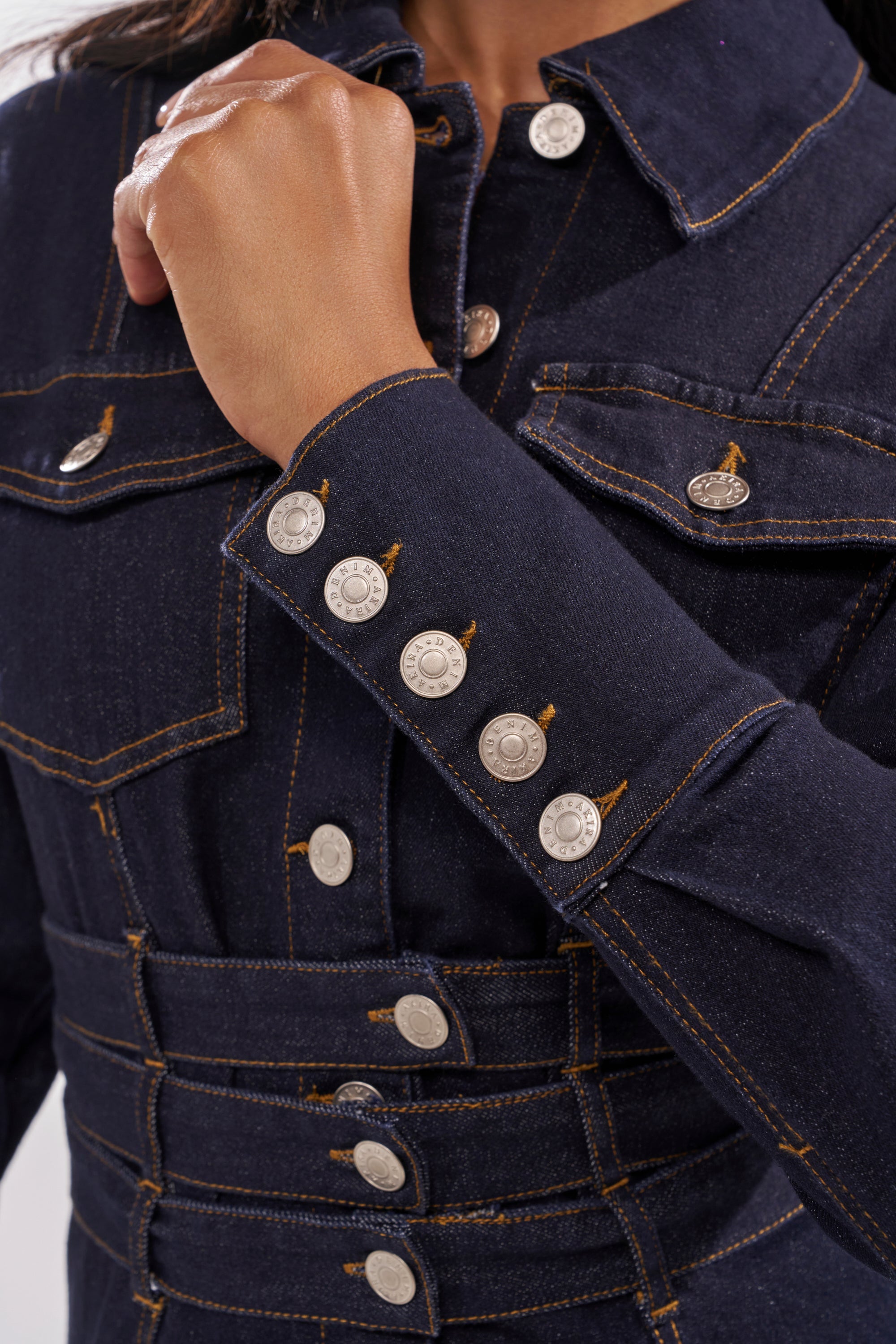 A close-up of a person wearing the CITY NIGHTS DENIM BUTTON DOWN, featuring silver buttons and orange stitching, with sleeve, chest, and waist details visible. The person's hand is raised near their collar. 6