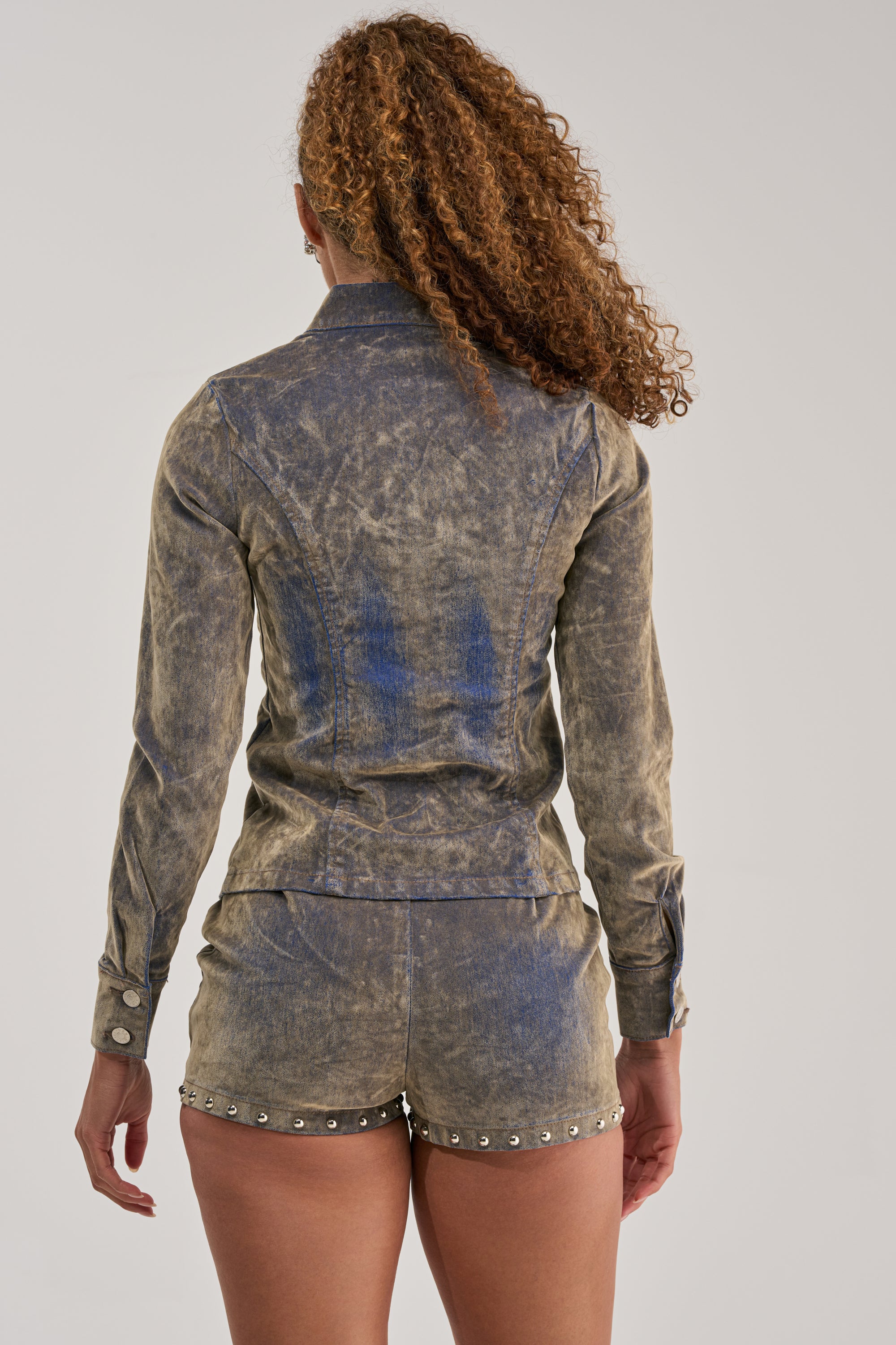 A person with curly hair wears the BURNT OUT ZIP UP BLOUSE, styled with matching studded shorts. Shown from the back against a plain, light background, their look highlights an effortlessly cool streetwear vibe.