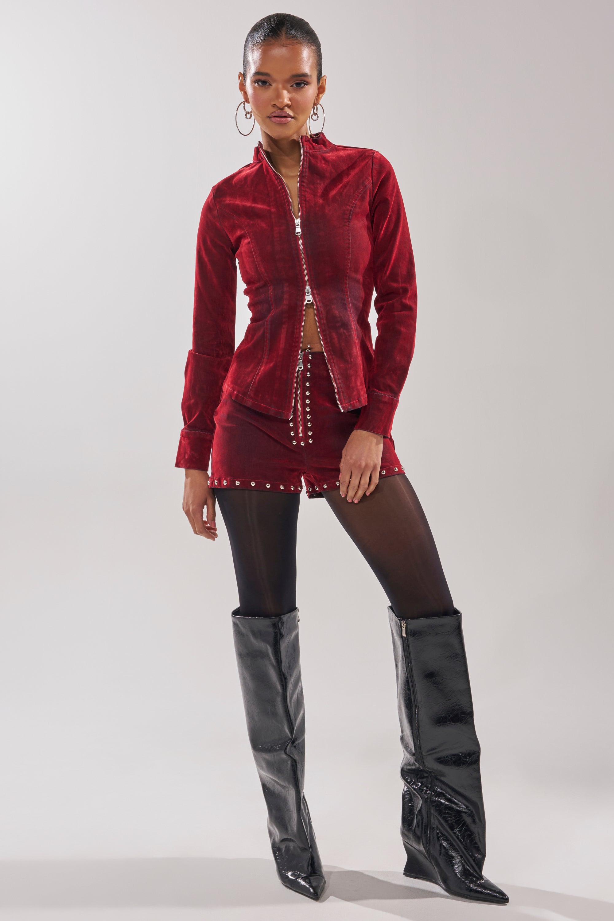 A woman poses against a plain light background in the RODEO VIBES VELVET DENIM STUDDED SHORT, paired with a red zip-up jacket, black tights, and tall slouchy black boots.