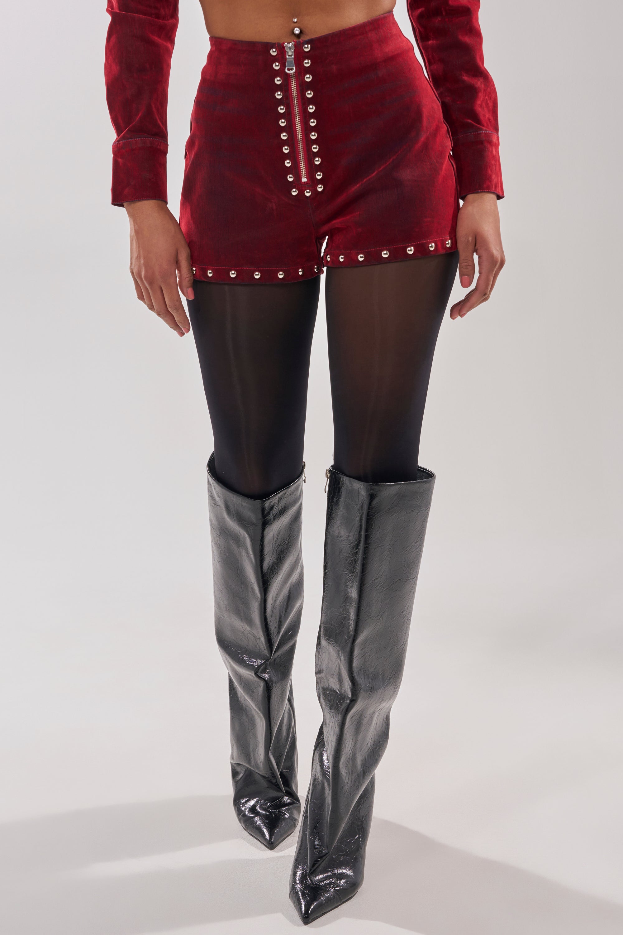Wearing the RODEO VIBES VELVET DENIM STUDDED SHORT, sheer black tights, and metallic silver knee-high boots. Only the lower body is visible; upper body and face are not shown.