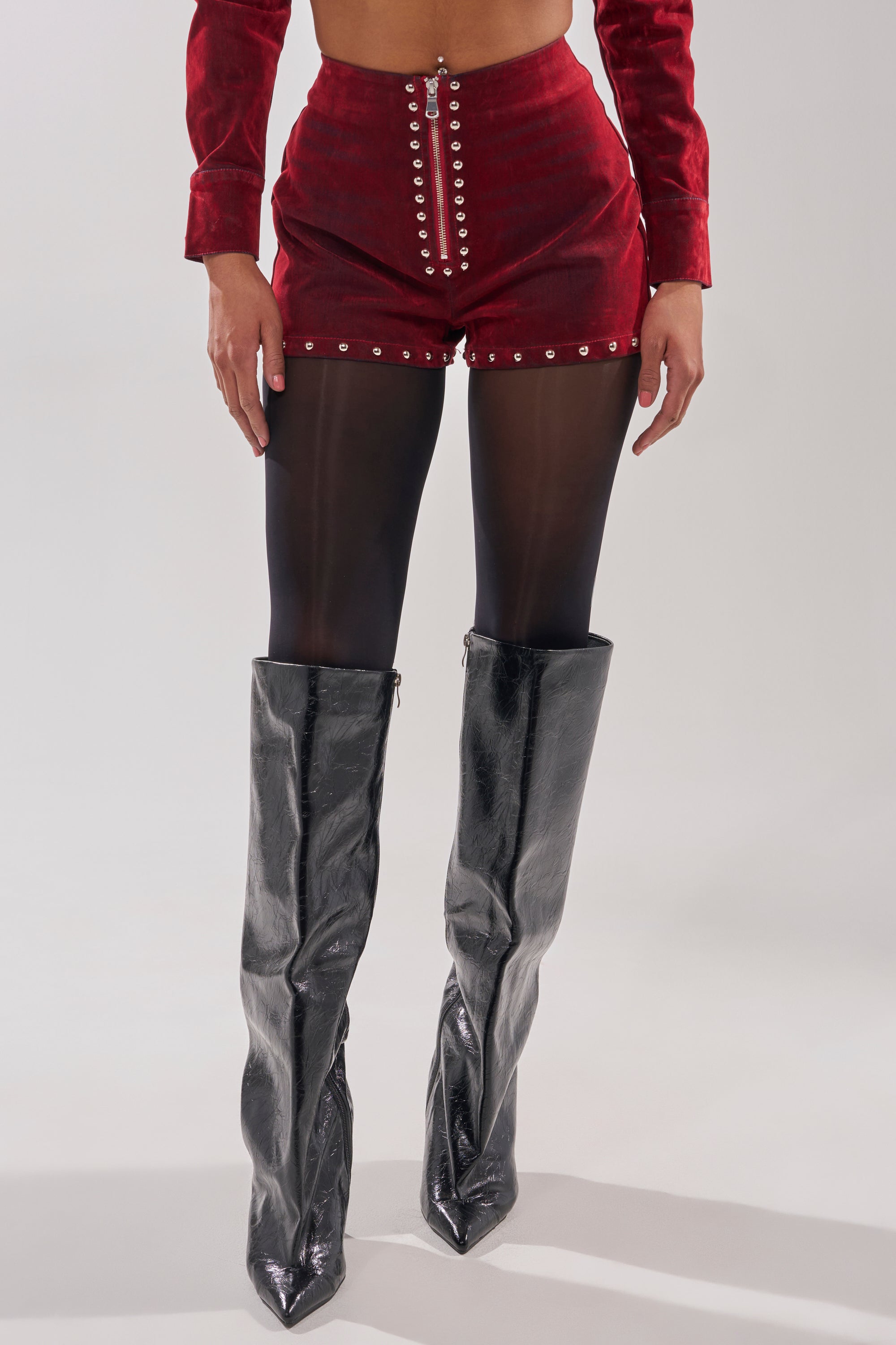 Wearing RODEO VIBES VELVET DENIM STUDDED SHORT with silver buttons, sheer black tights, and shiny black knee-high boots, a person stands against a plain light background. Their face is not visible.